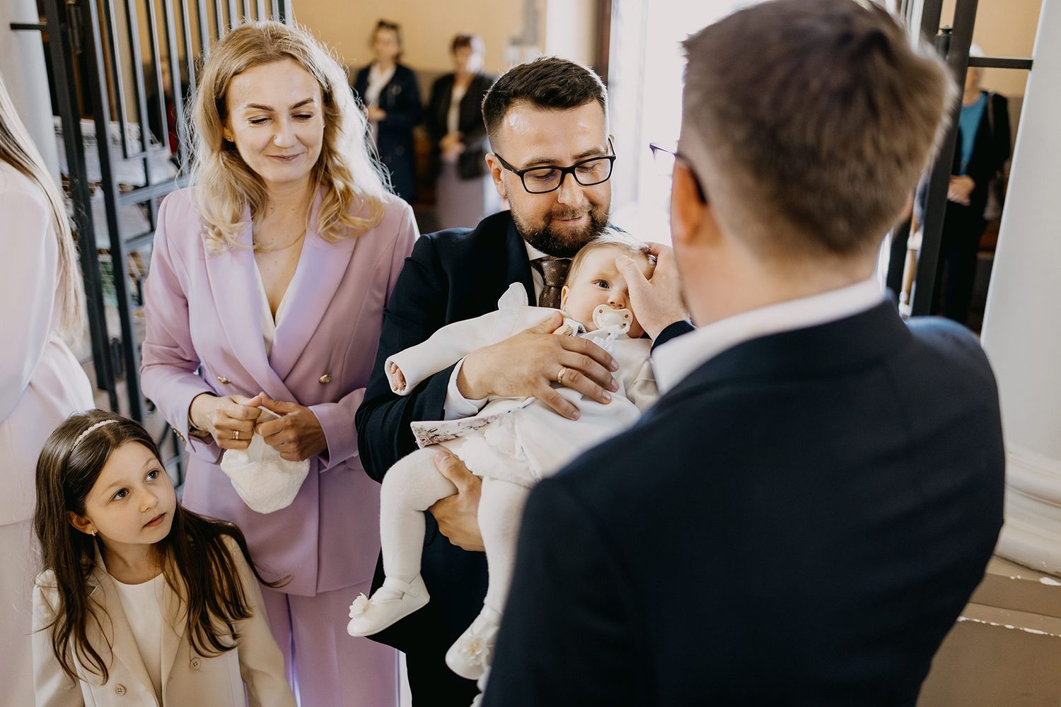 A family with a young girl and baby in white christening gown greets a guest indoors.