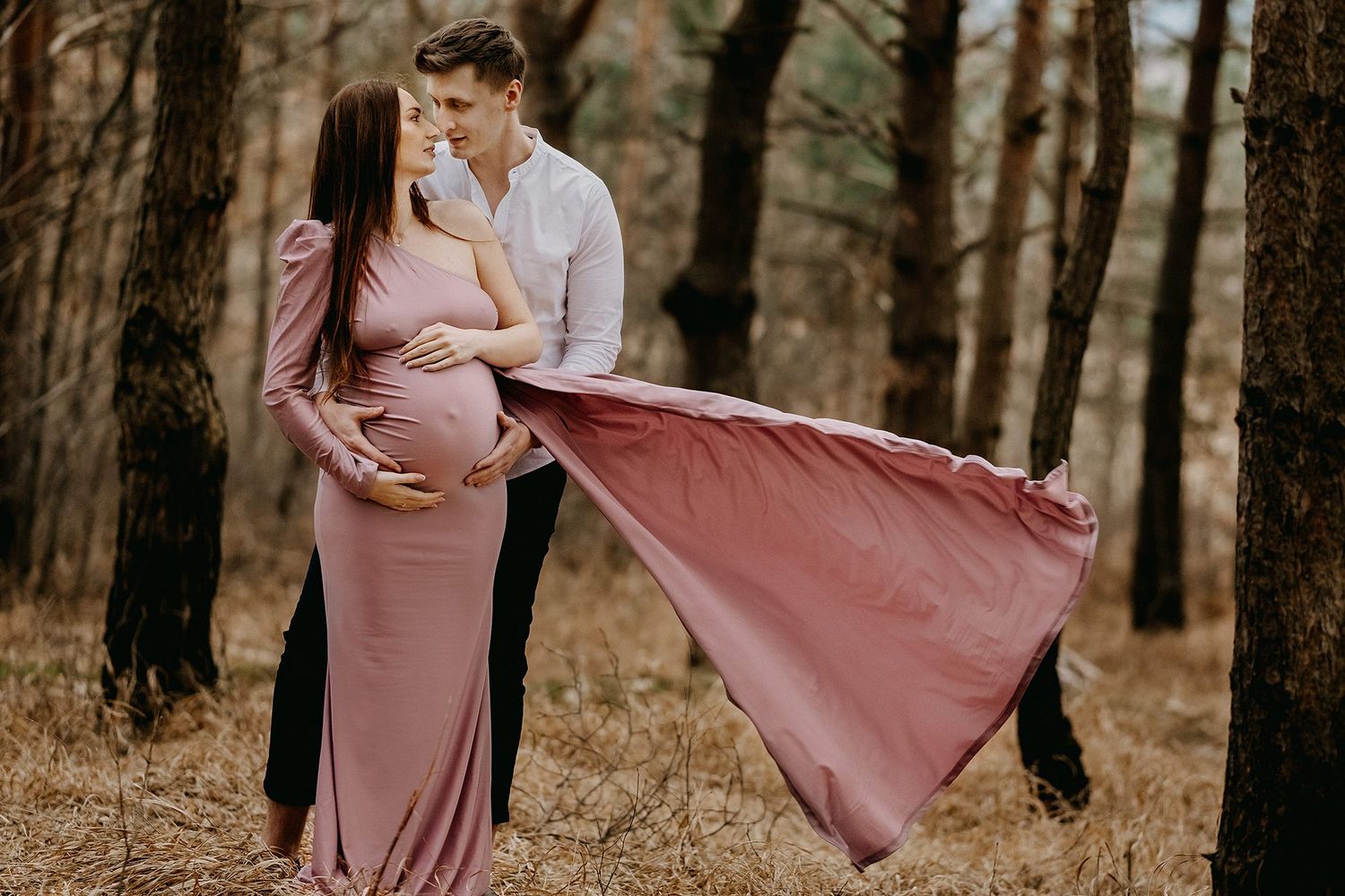 Couple sharing romantic moment in pine forest, pregnant woman's pink gown flowing in wind.