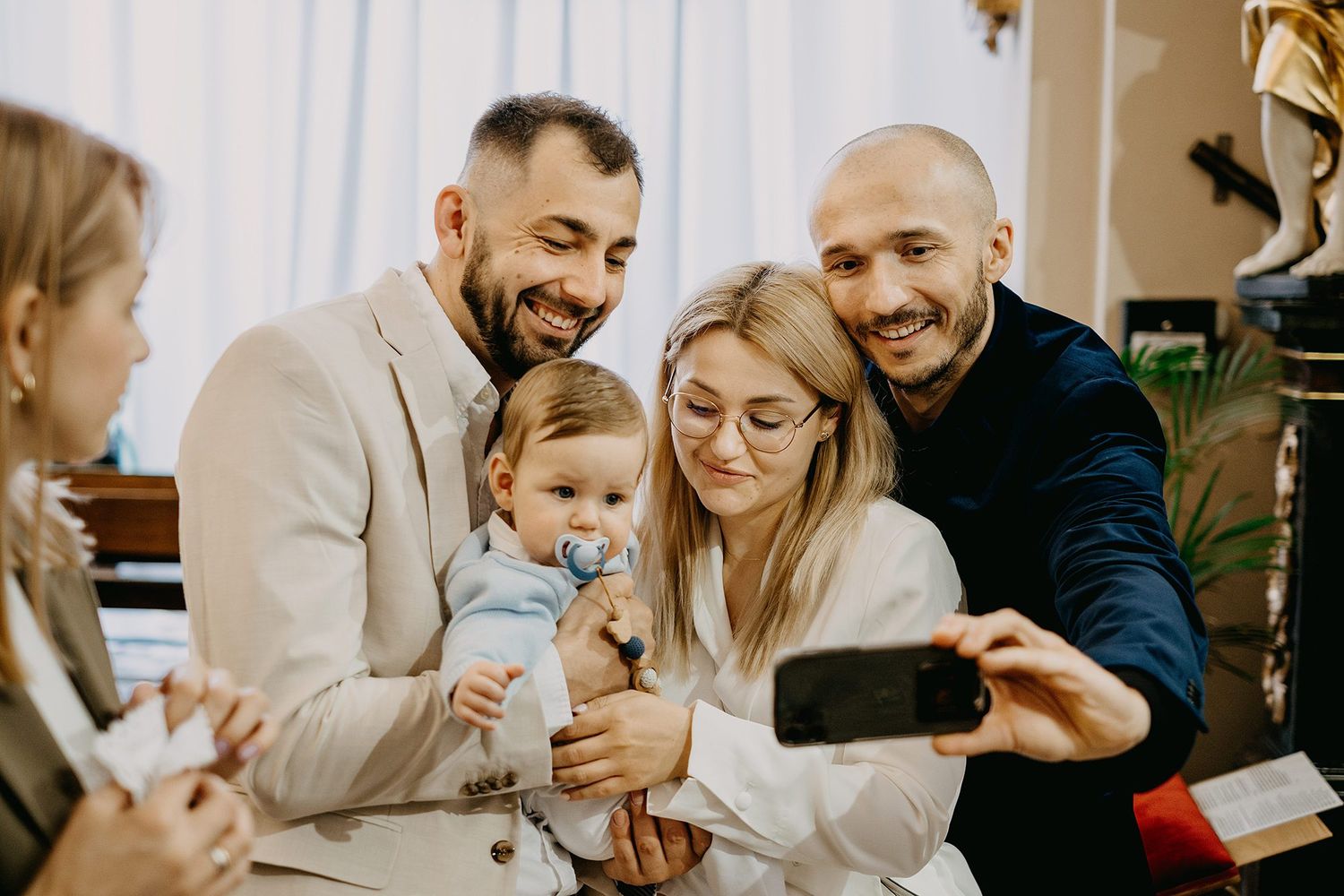 Parents and godfather take selfie with baby using pacifier at baptism celebration gathering.