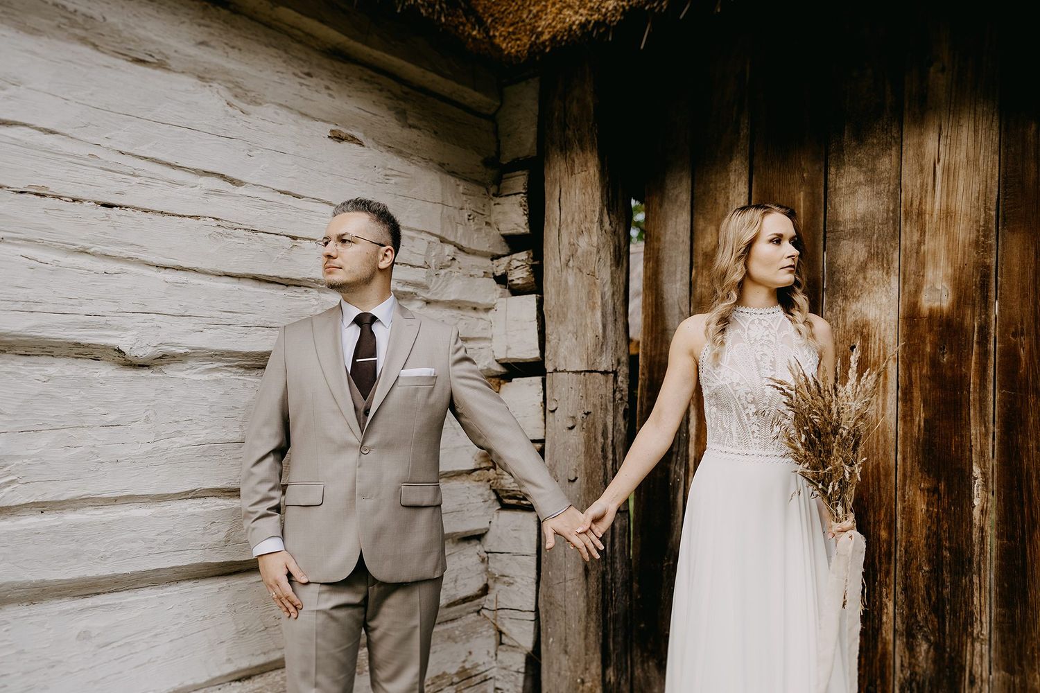 Bride and groom holding hands standing apart by a rustic wooden barn wall in bohemian attire.