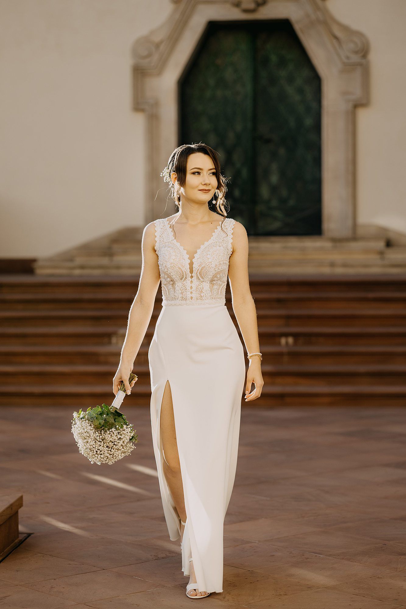 Elegant bride in a sleek white gown with lace bodice walking with a bouquet near a stone building.
