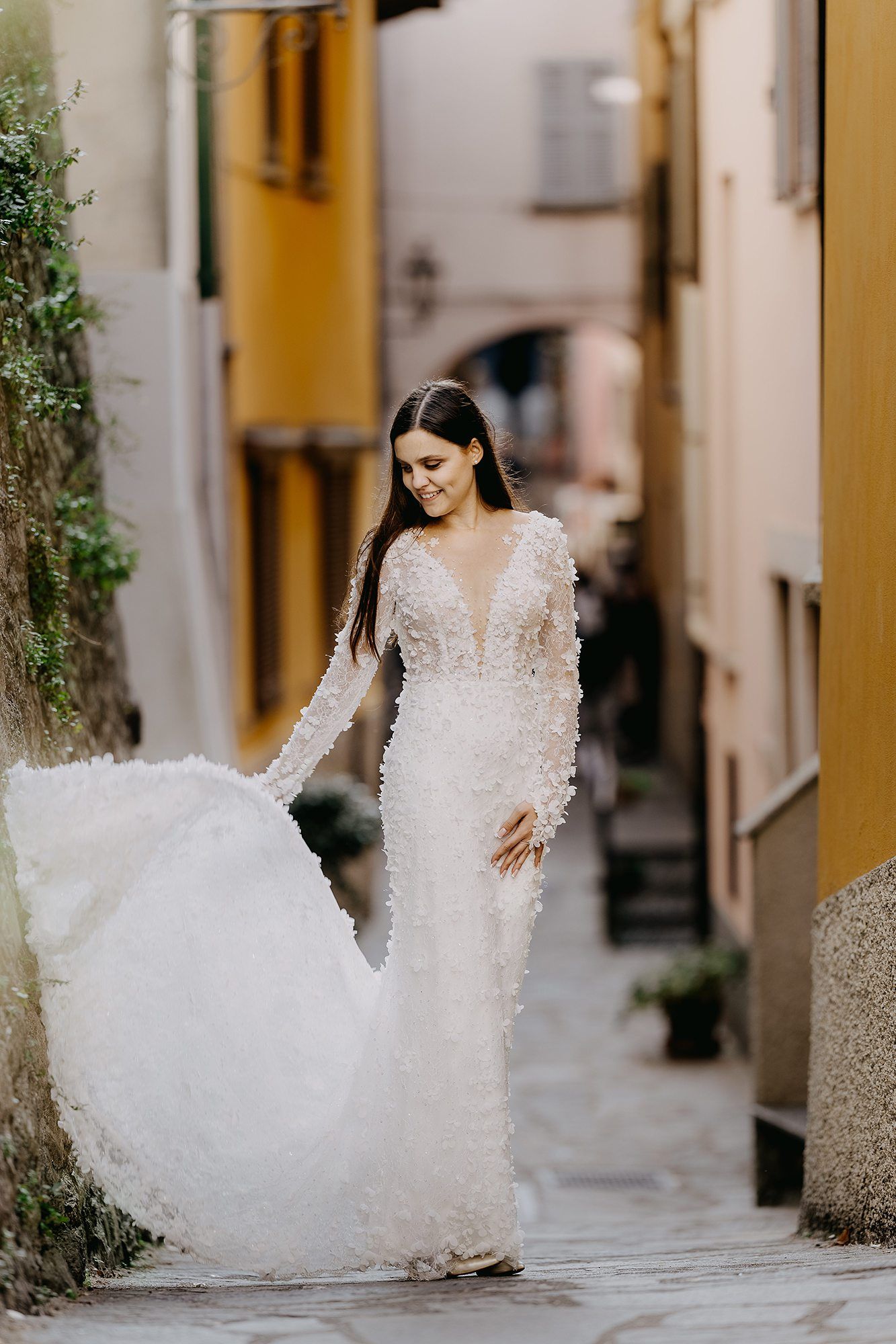 Bride in floral long-sleeve white gown walks through a charming narrow Italian village alleyway.