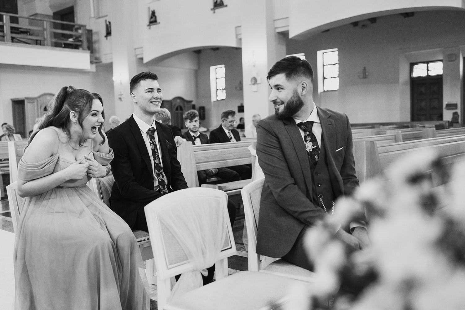 Black and white photo of wedding guests laughing and chatting inside a bright church before ceremony.