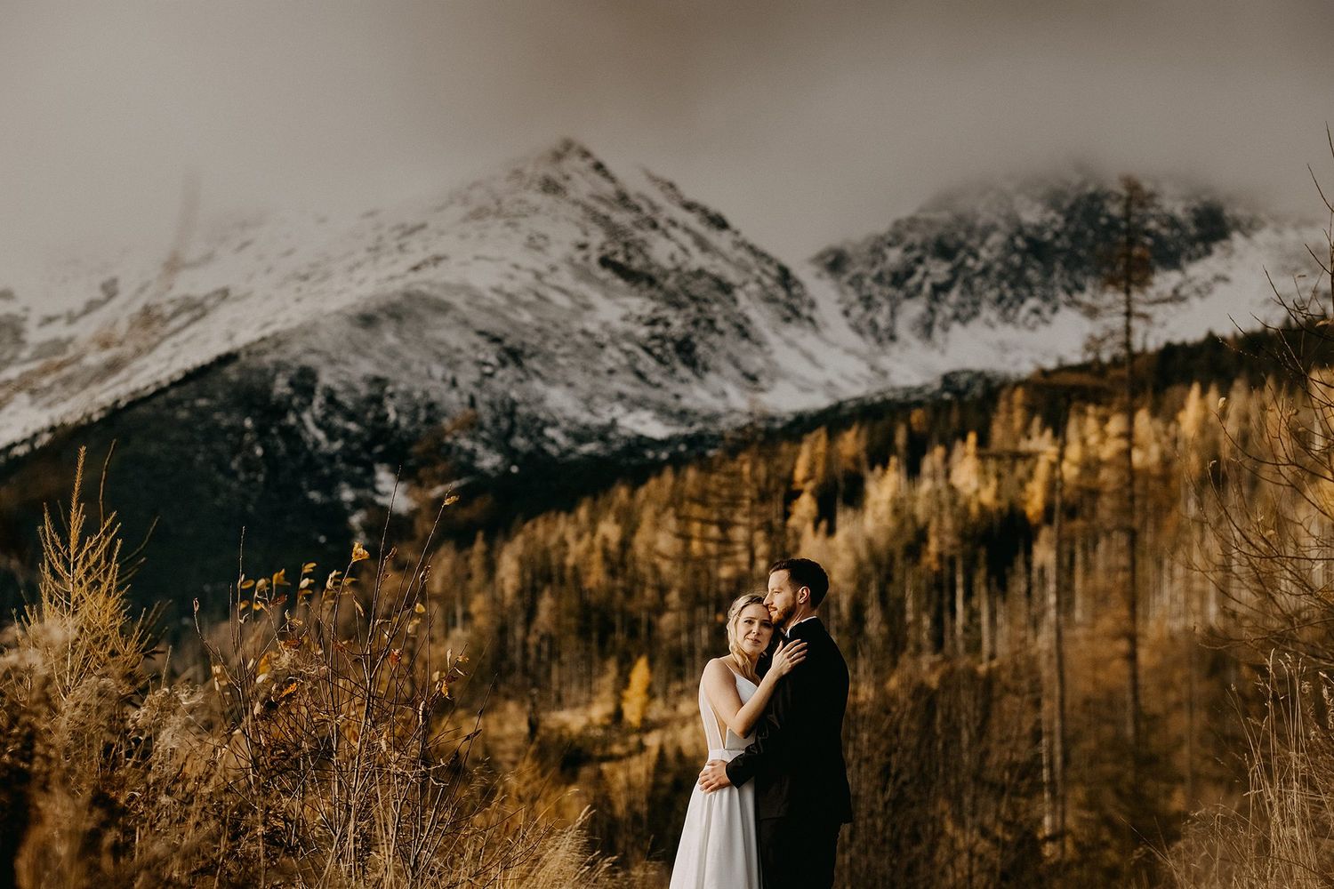 Bride and groom embrace in an autumn mountain landscape with snow-capped peaks behind them.