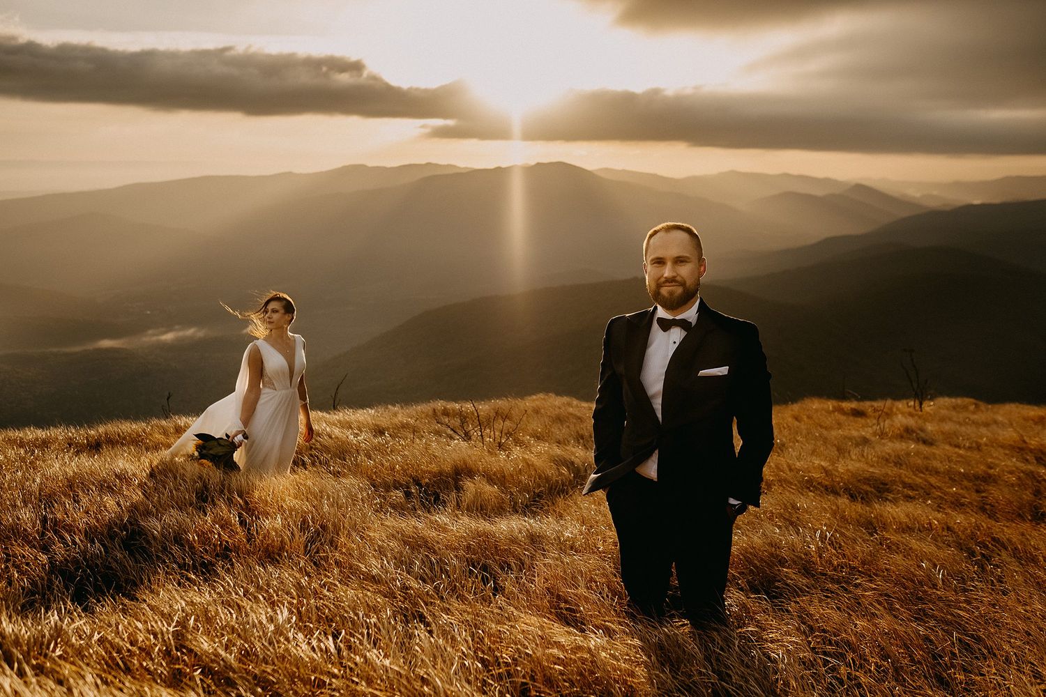 A groom stands confidently in a tuxedo as his bride poses behind him on a golden mountain.