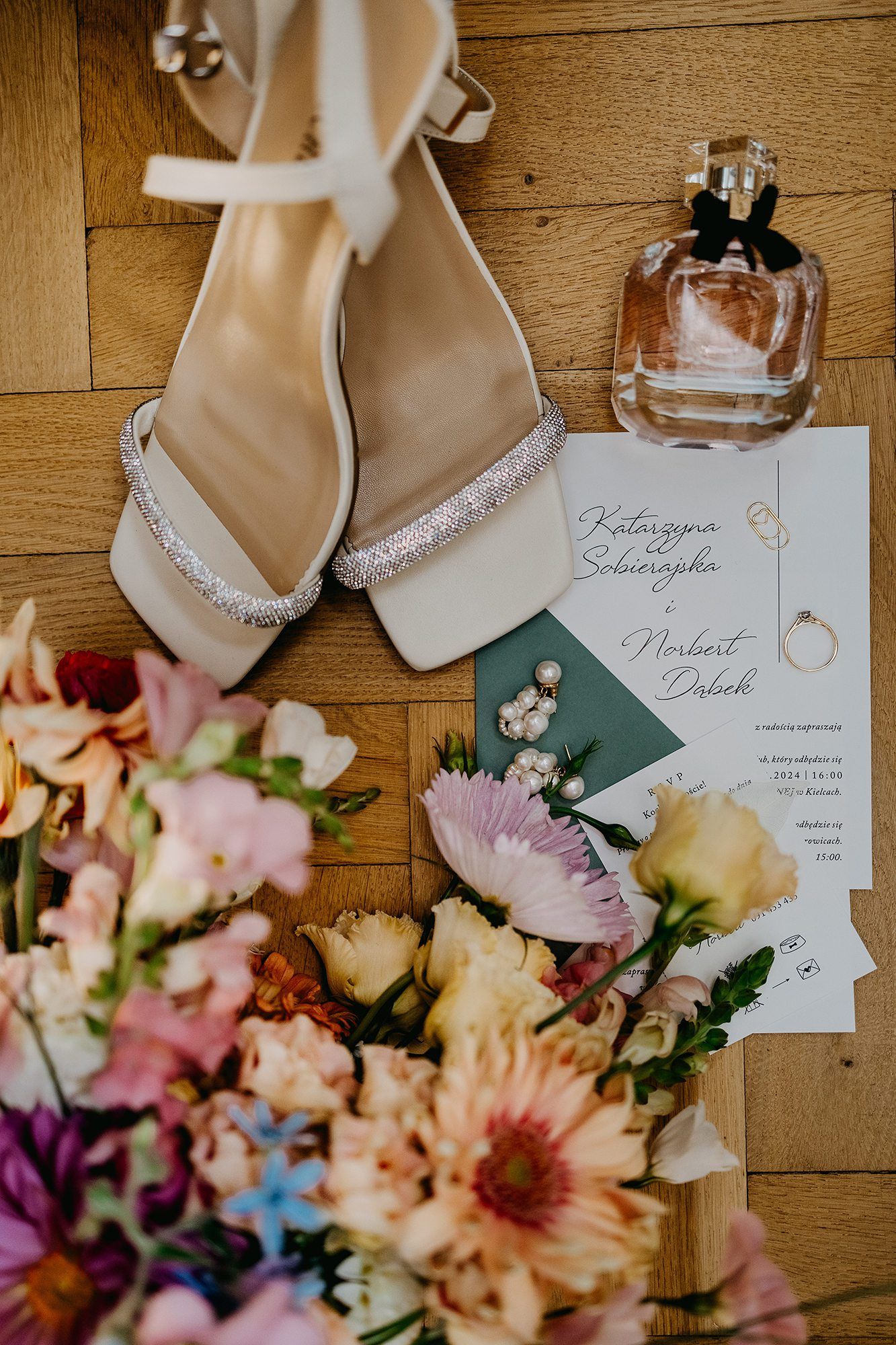 Bridal flat lay with glitter heels, wedding invitation, pearl earrings, perfume, and colorful bouquet.