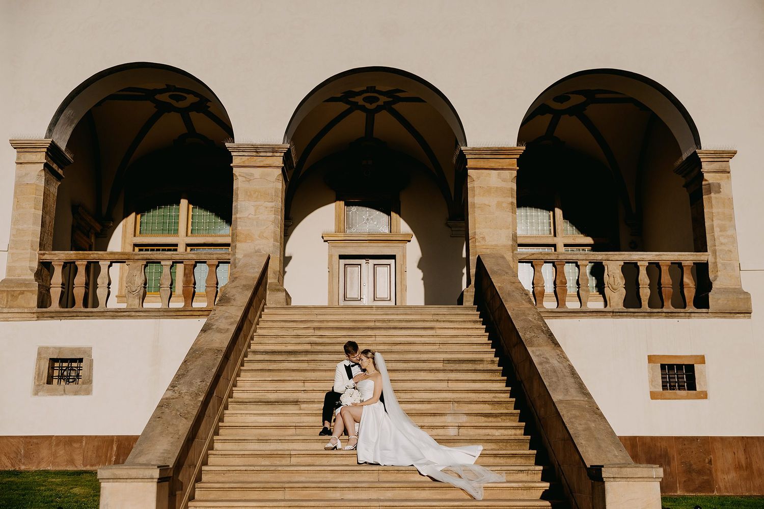 Bride and groom sitting romantically on grand stone staircase of elegant classical building with arched colonnade.