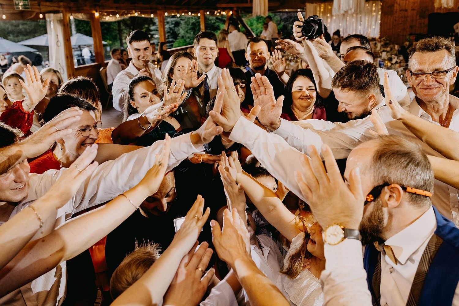 Large group of wedding guests reaching hands together in a joyful huddle celebration at an indoor reception venue.