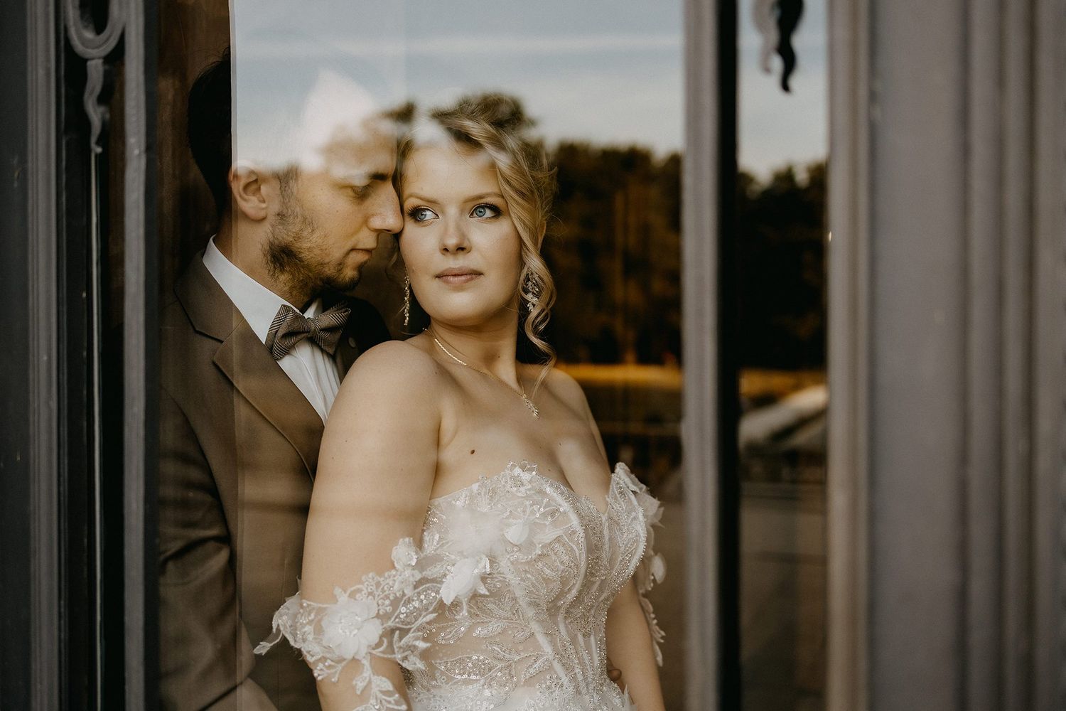Elegant bride in floral wedding gown standing by glass door with groom reflected behind her.