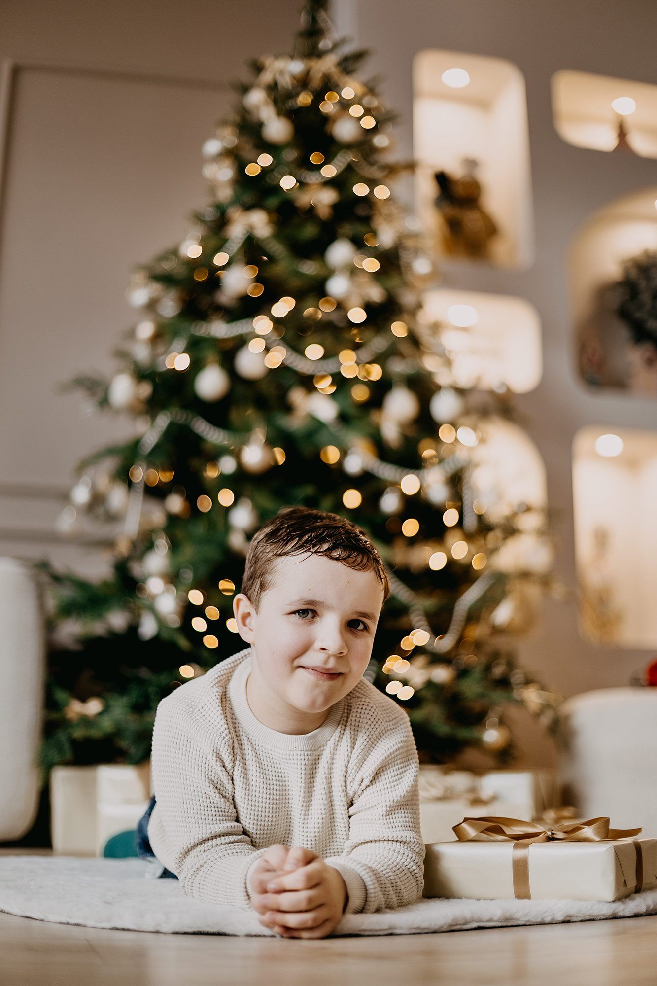 A young boy in a cream sweater lies on the floor smiling in front of a decorated Christmas tree with gifts.