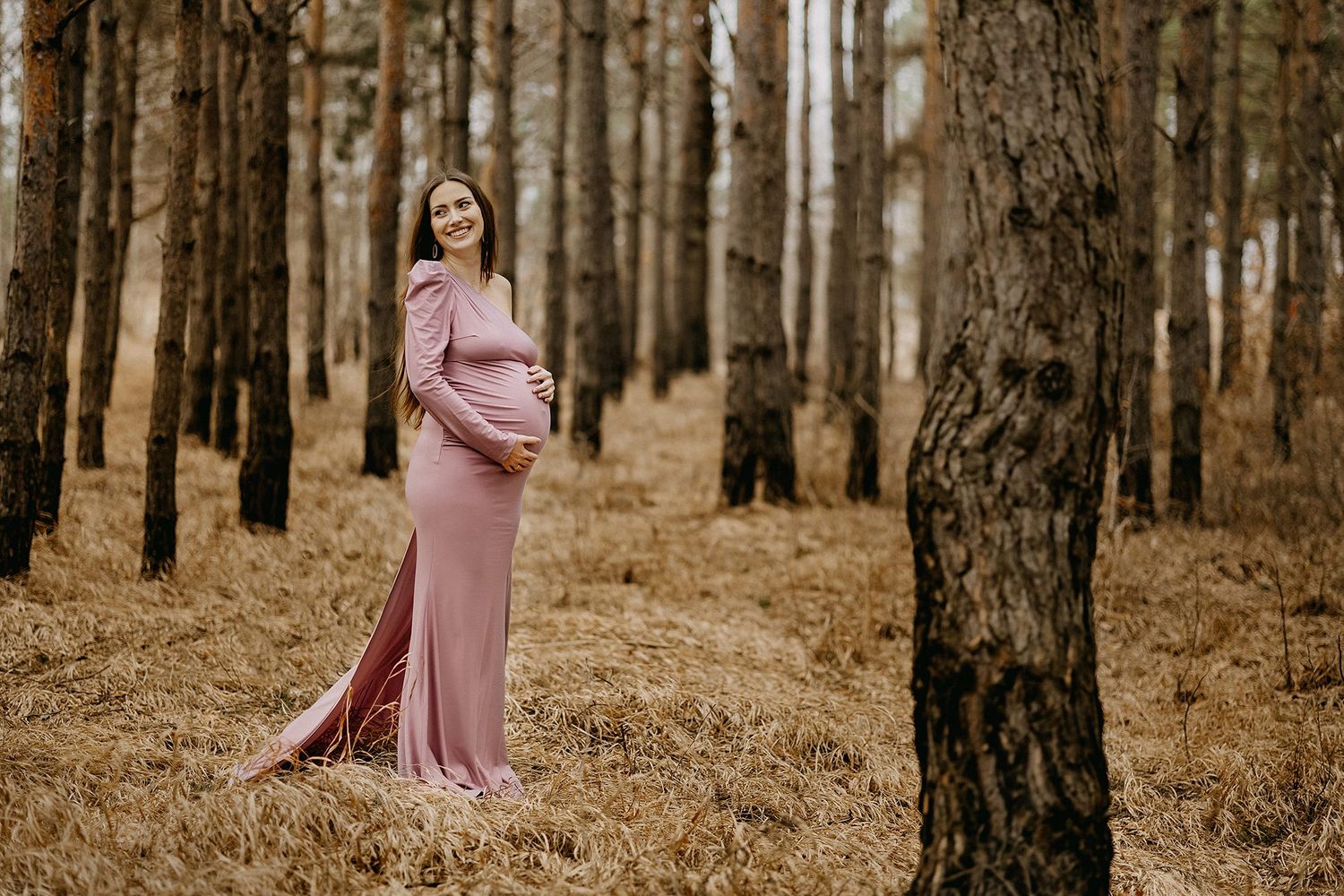 Smiling pregnant woman in elegant pink gown posing in golden autumn pine forest.