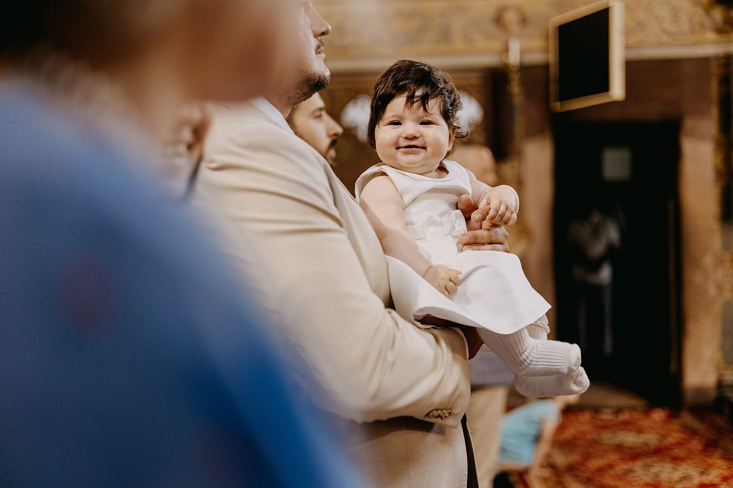 Smiling baby in white dress being held up by man in beige suit at ceremony.