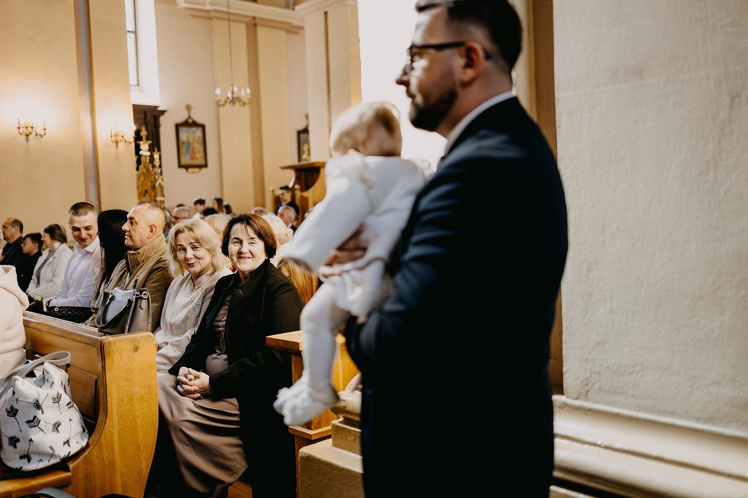 Father holds baby in white christening outfit while smiling family members sit in church pews.