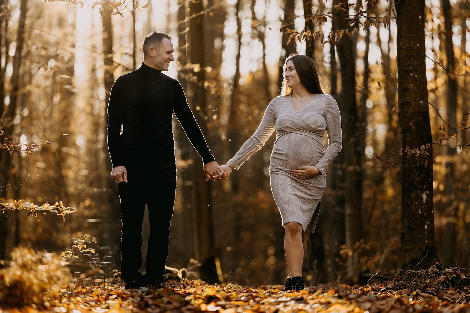 Pregnant couple holding hands in autumn forest, woman in gray dress cradling baby bump, golden fall foliage background.