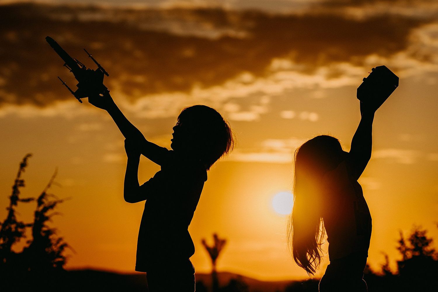 Silhouette of two children playing with toy airplane against a dramatic orange sunset sky.