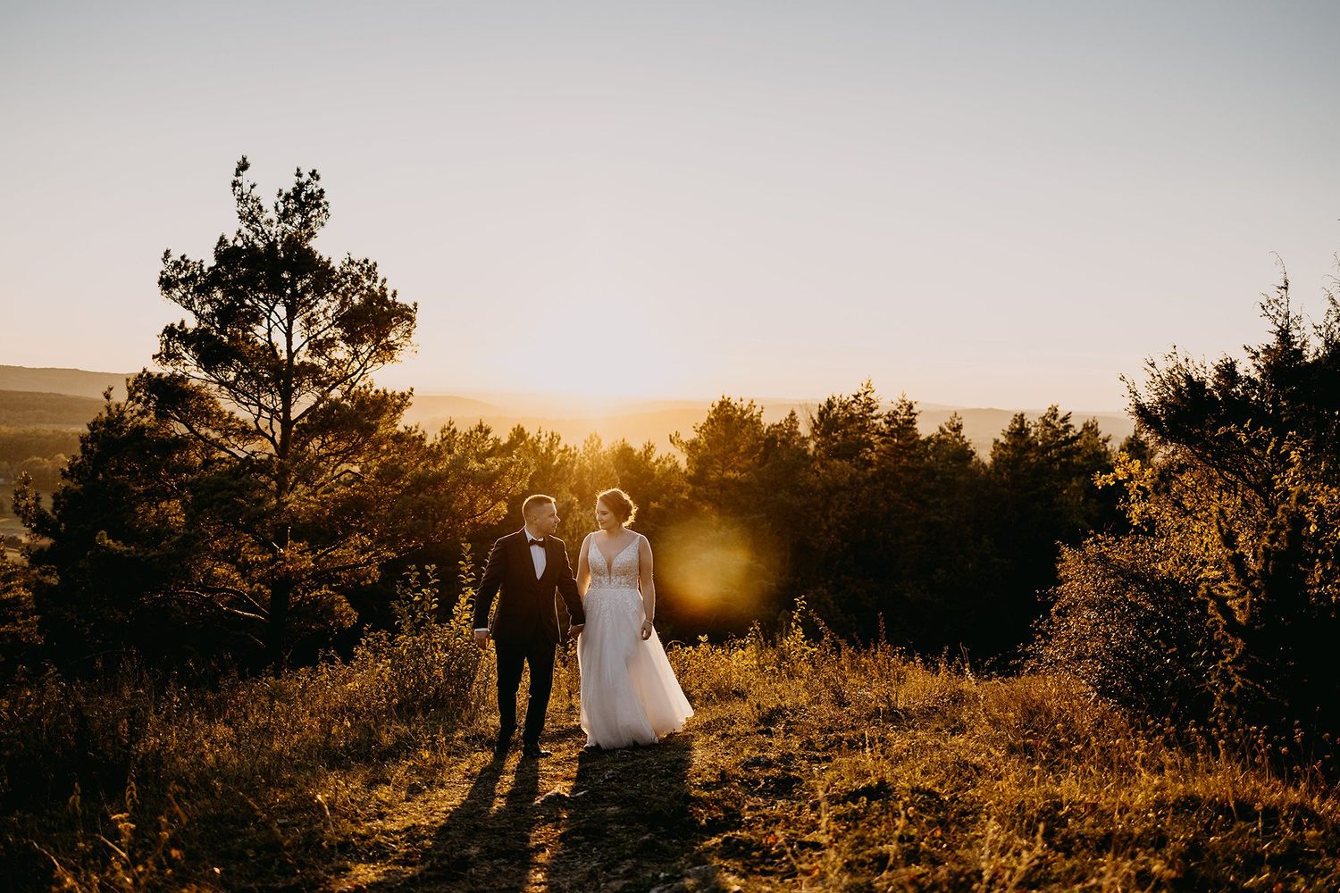 Bride and groom walking through golden meadow at sunset with pine trees glowing in warm light.