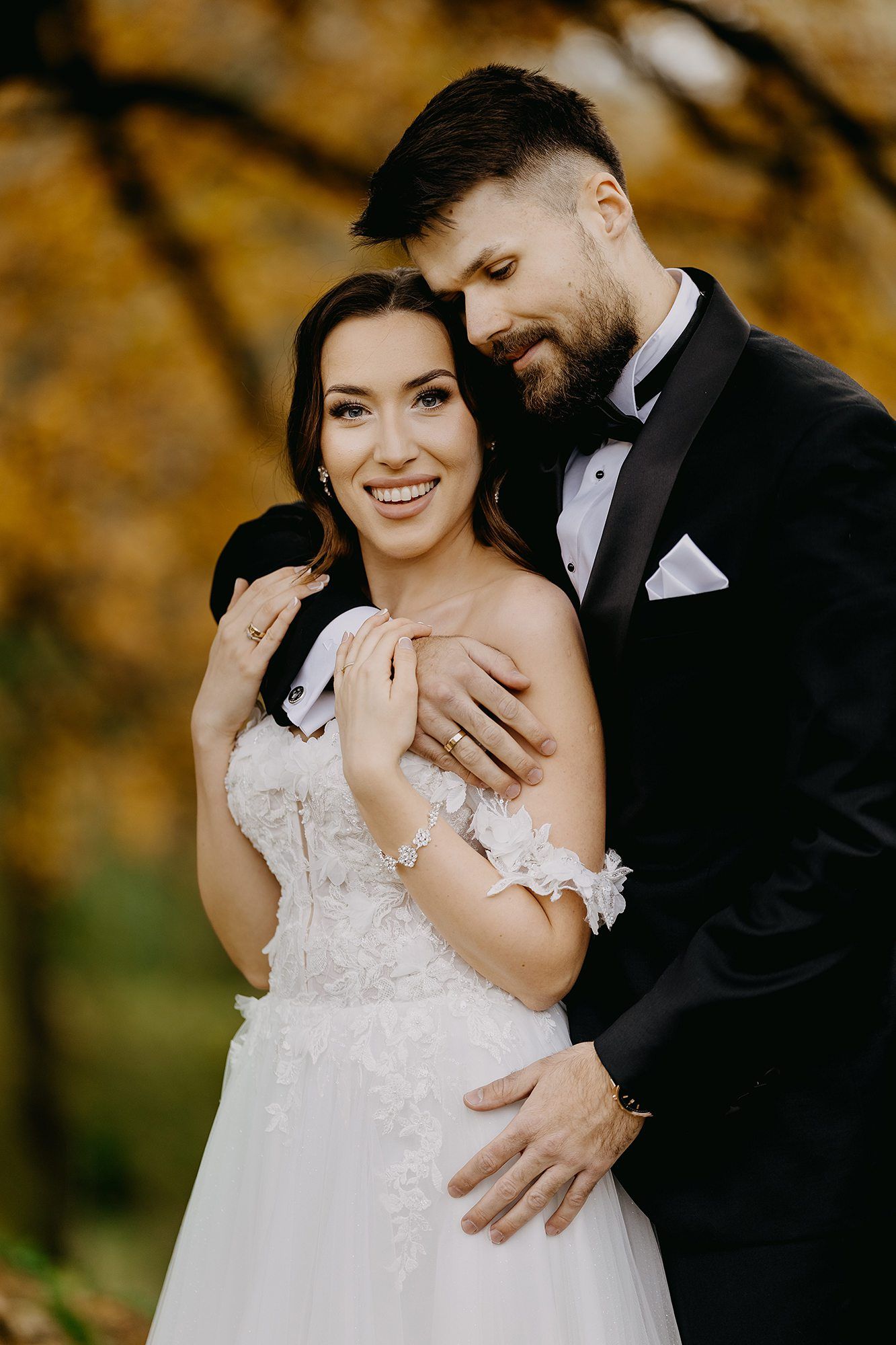 Smiling bride in lace gown embraced by bearded groom in black tuxedo against autumn foliage backdrop.