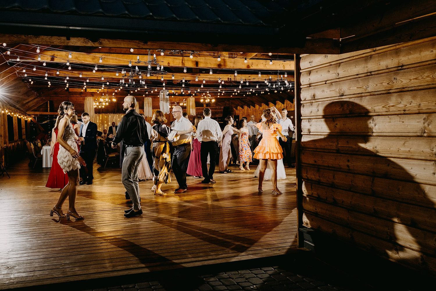 Wedding guests dancing in a warmly lit rustic barn venue with string lights overhead.