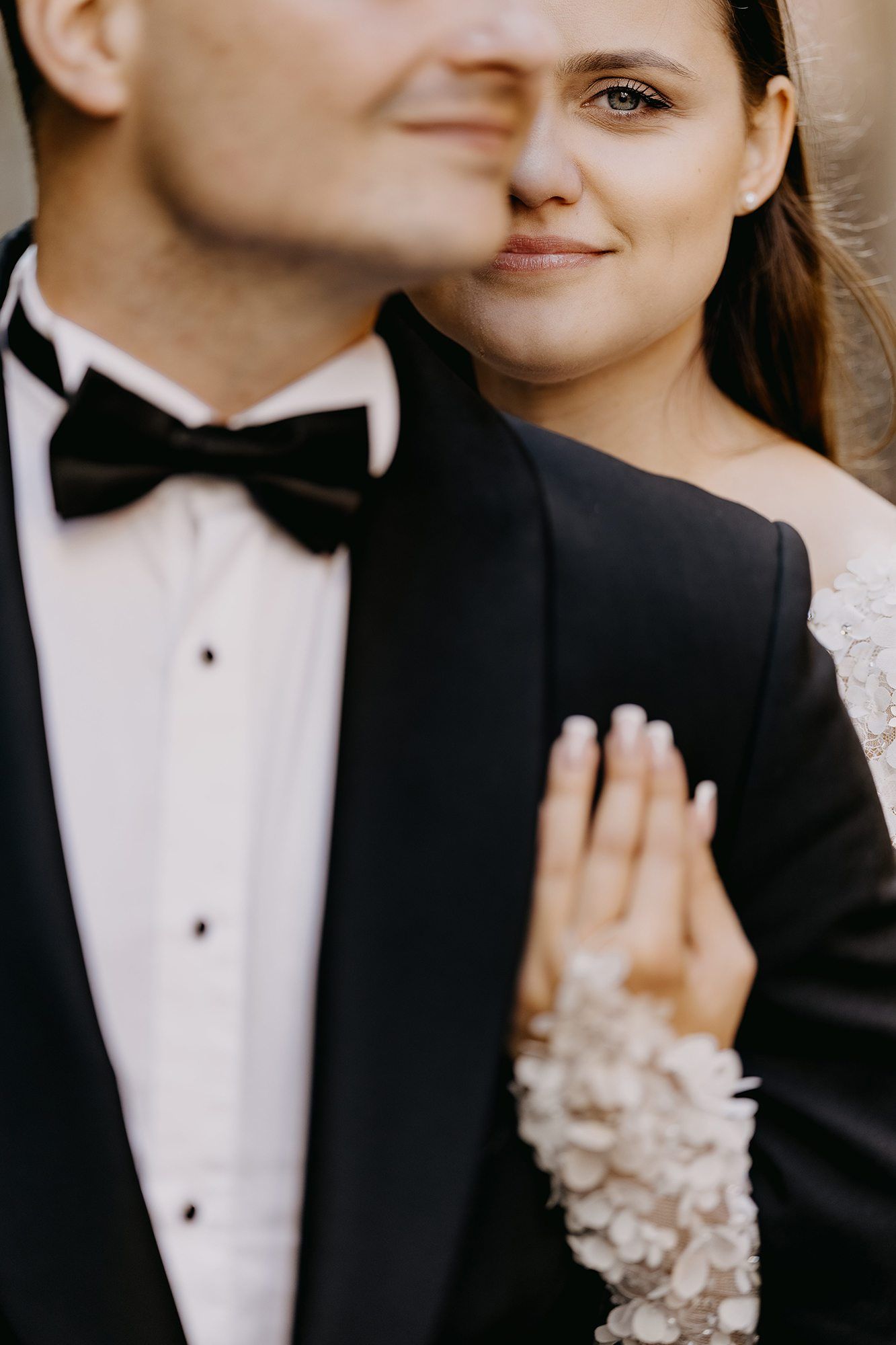 Bride in floral lace gown rests hand on groom's black tuxedo lapel during intimate wedding portrait.