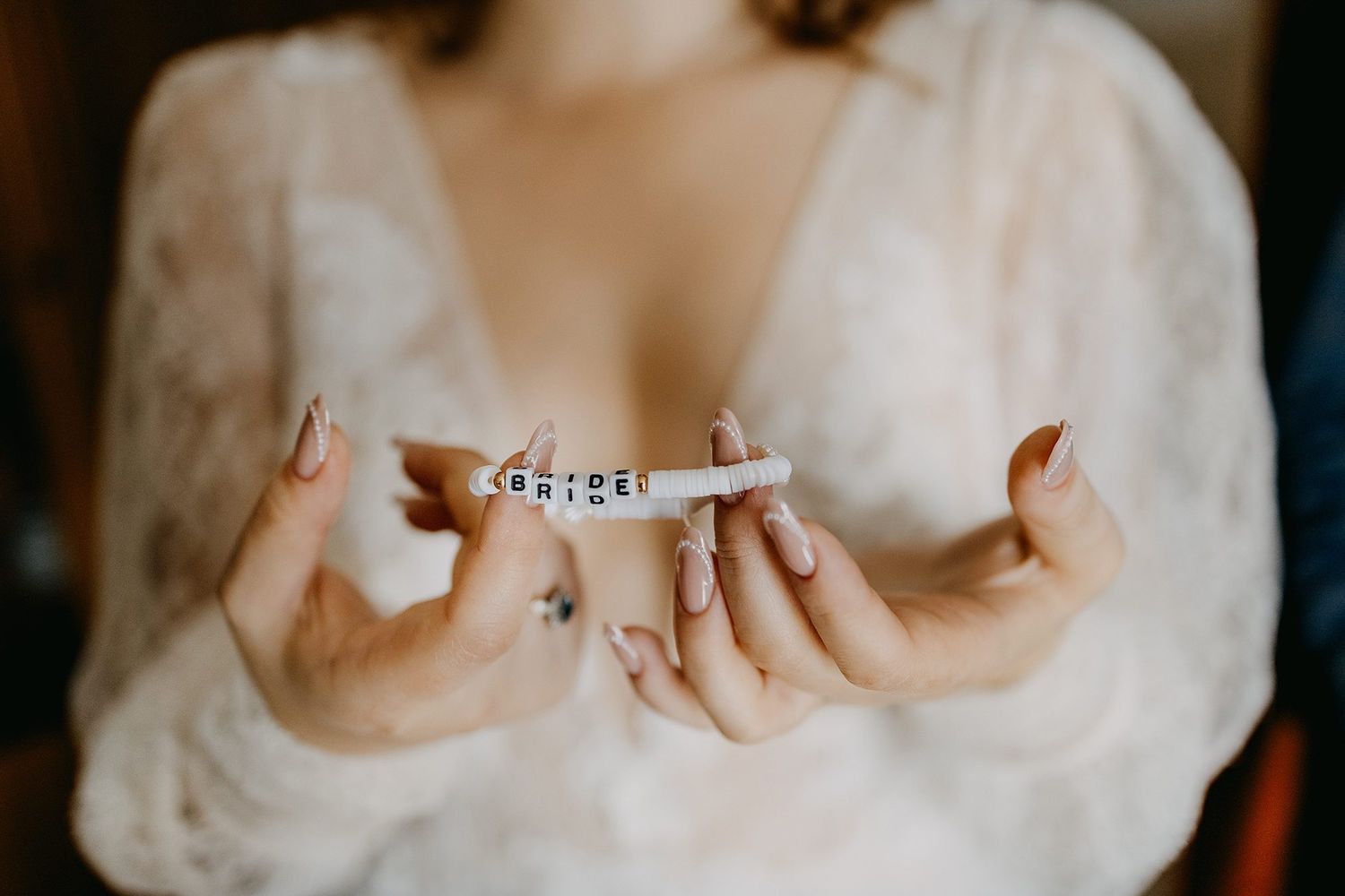 Bride in lace robe holding a beaded bracelet spelling BRIDE with elegant manicured nails.