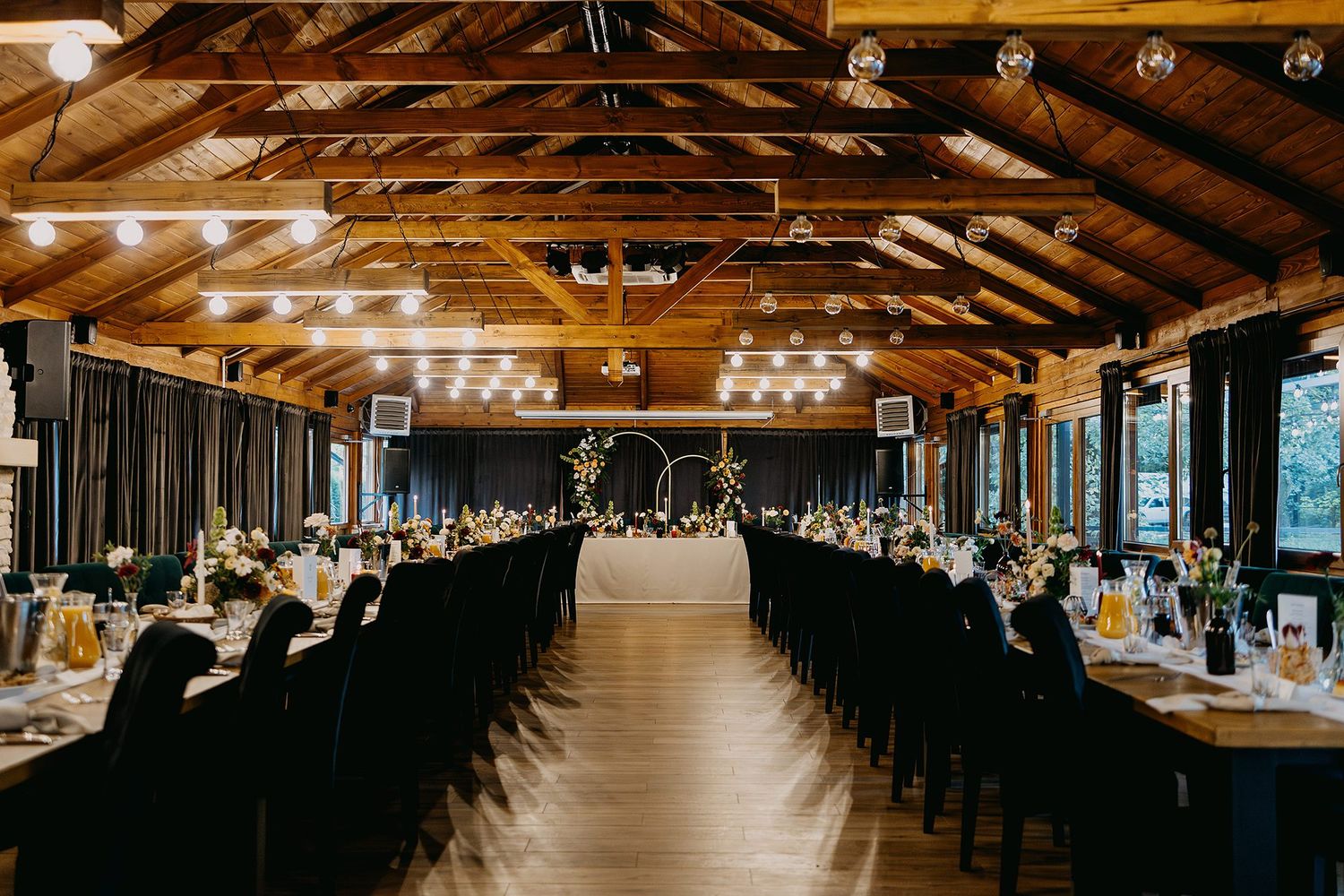 Rustic wedding reception hall with long banquet tables, string lights, and a floral arch centerpiece.