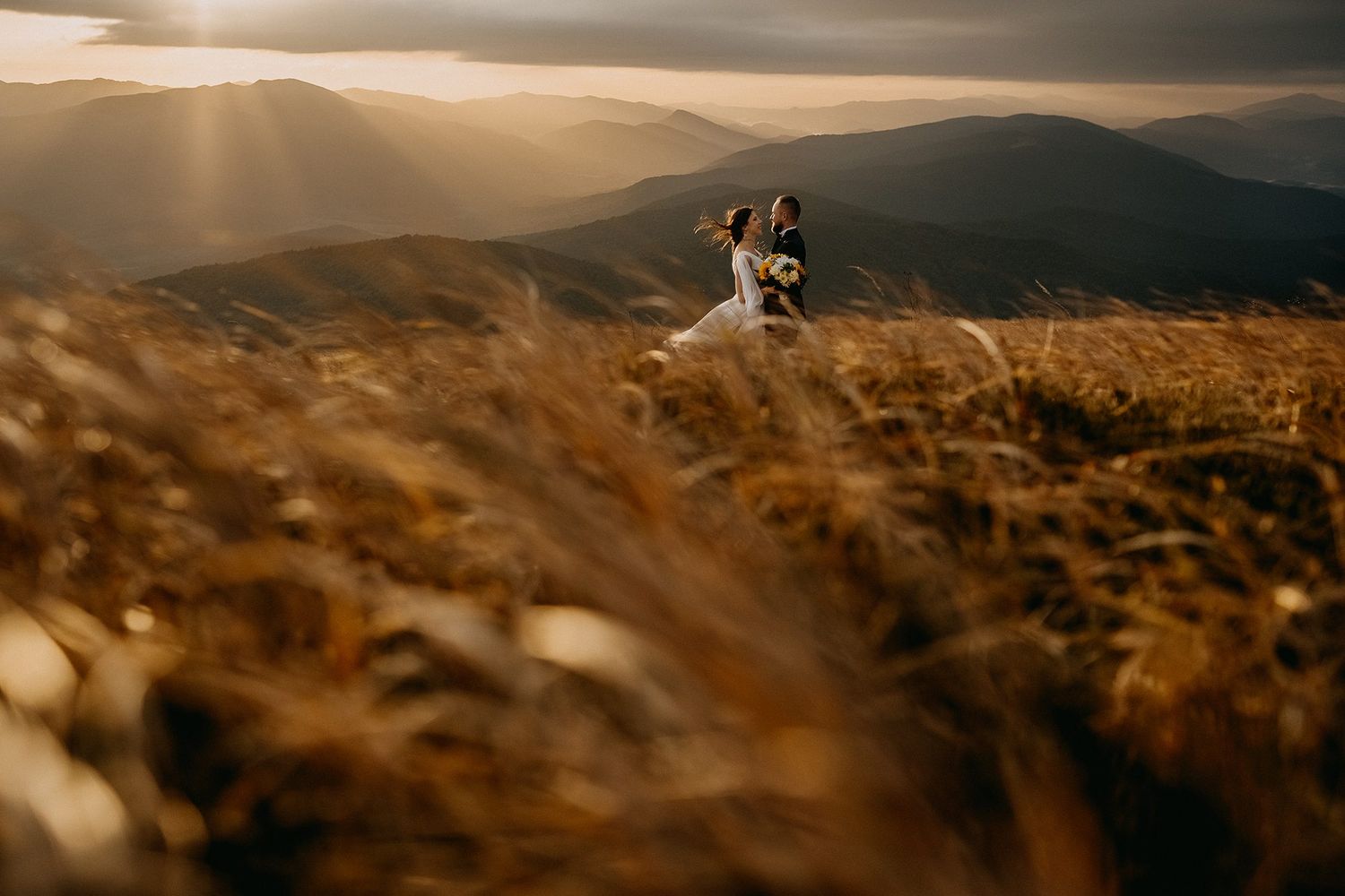 A wedding couple embraces in a windswept golden meadow with dramatic mountain sunset backdrop.