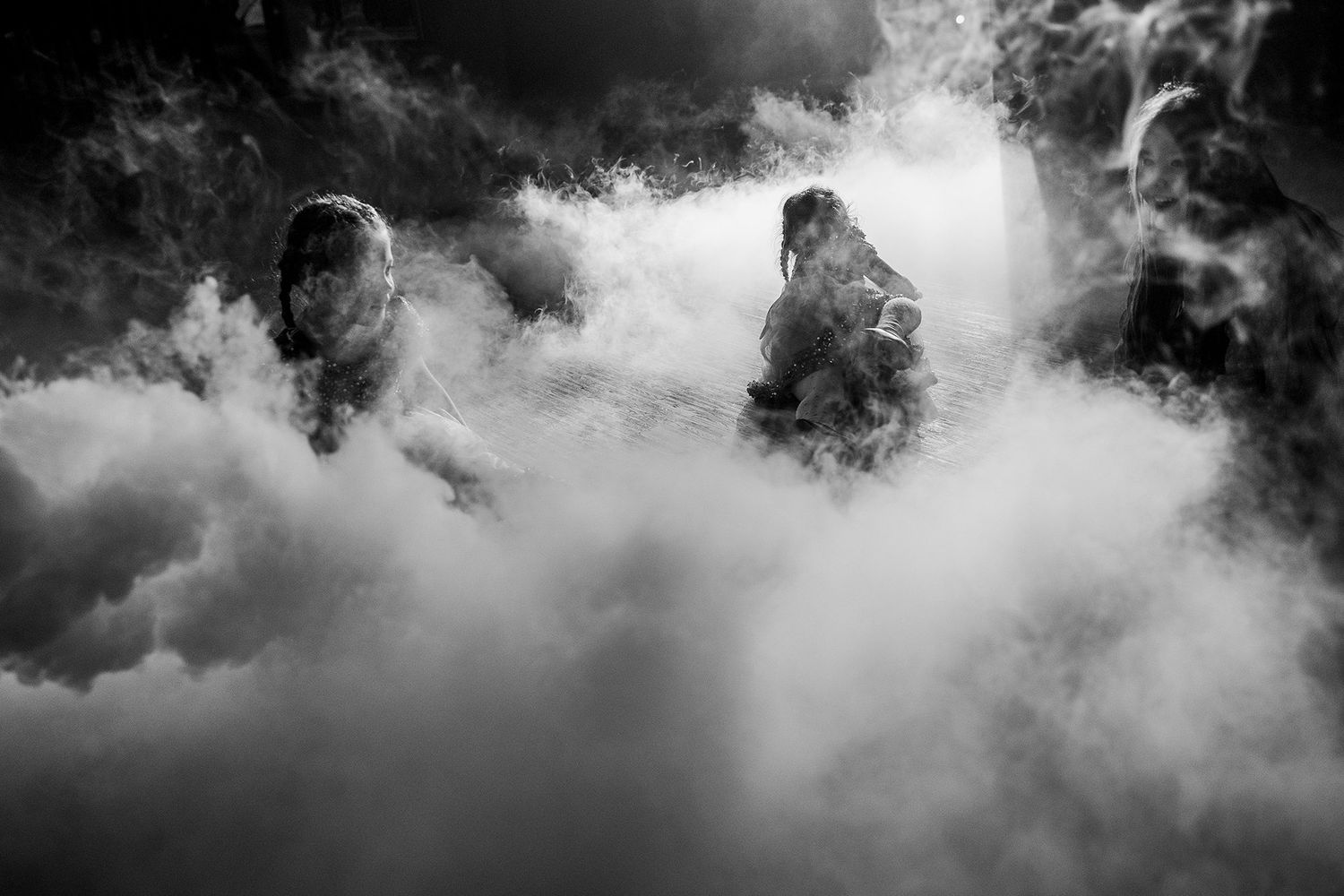Dramatic black and white photo of people surrounded by billowing smoke clouds at an outdoor event.