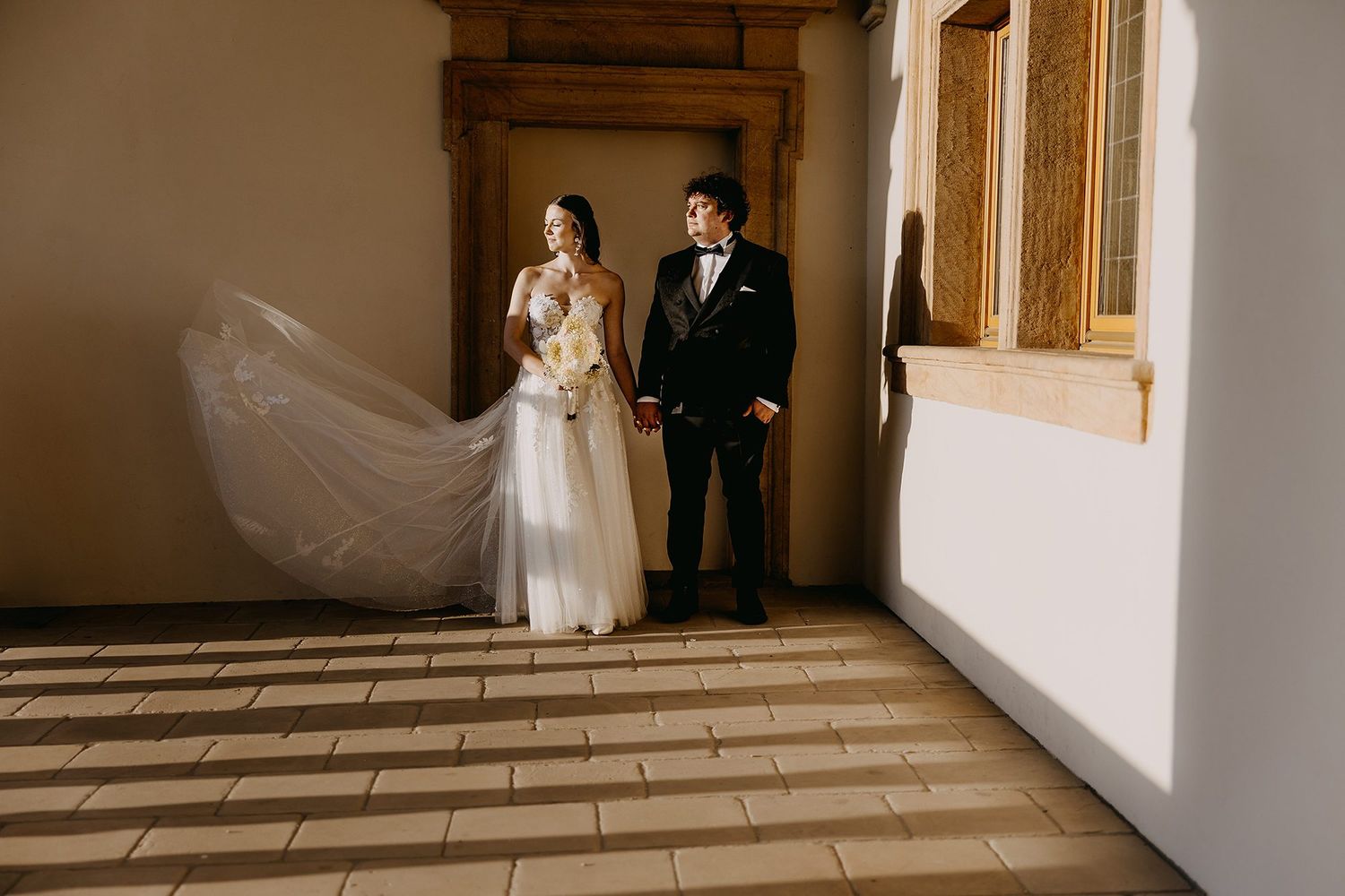 Bride and groom standing together in elegant wedding attire with veil flowing in a sunlit architectural hallway.
