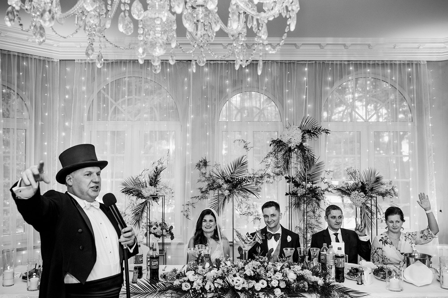 Wedding MC in top hat entertains guests at elegantly decorated reception table with floral arrangements.