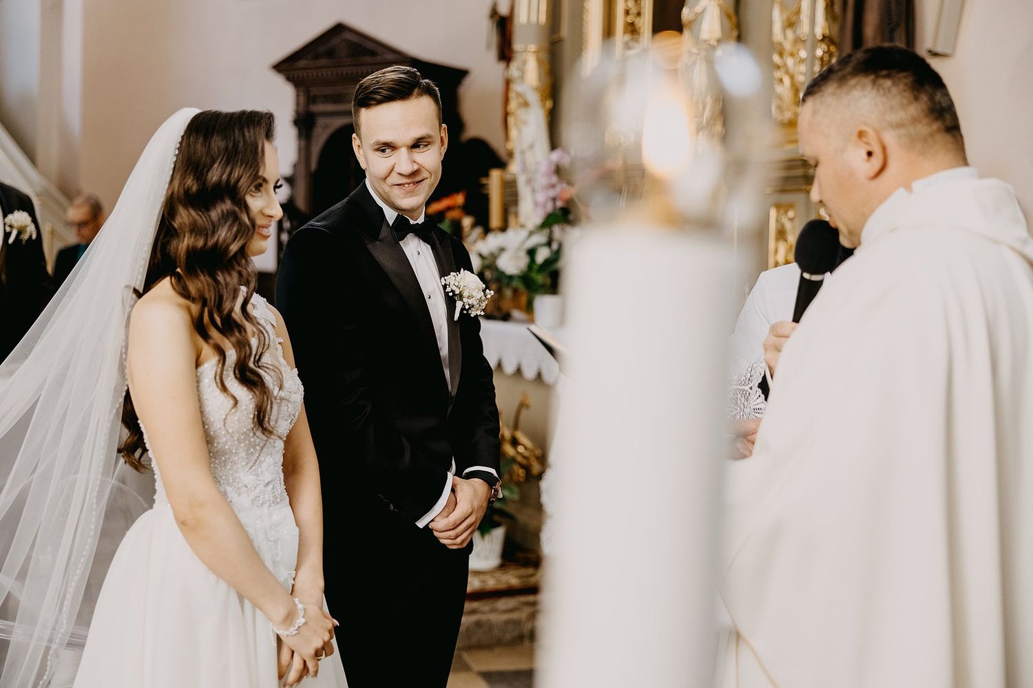 Bride and groom stand before priest during traditional church wedding ceremony, groom smiling warmly.