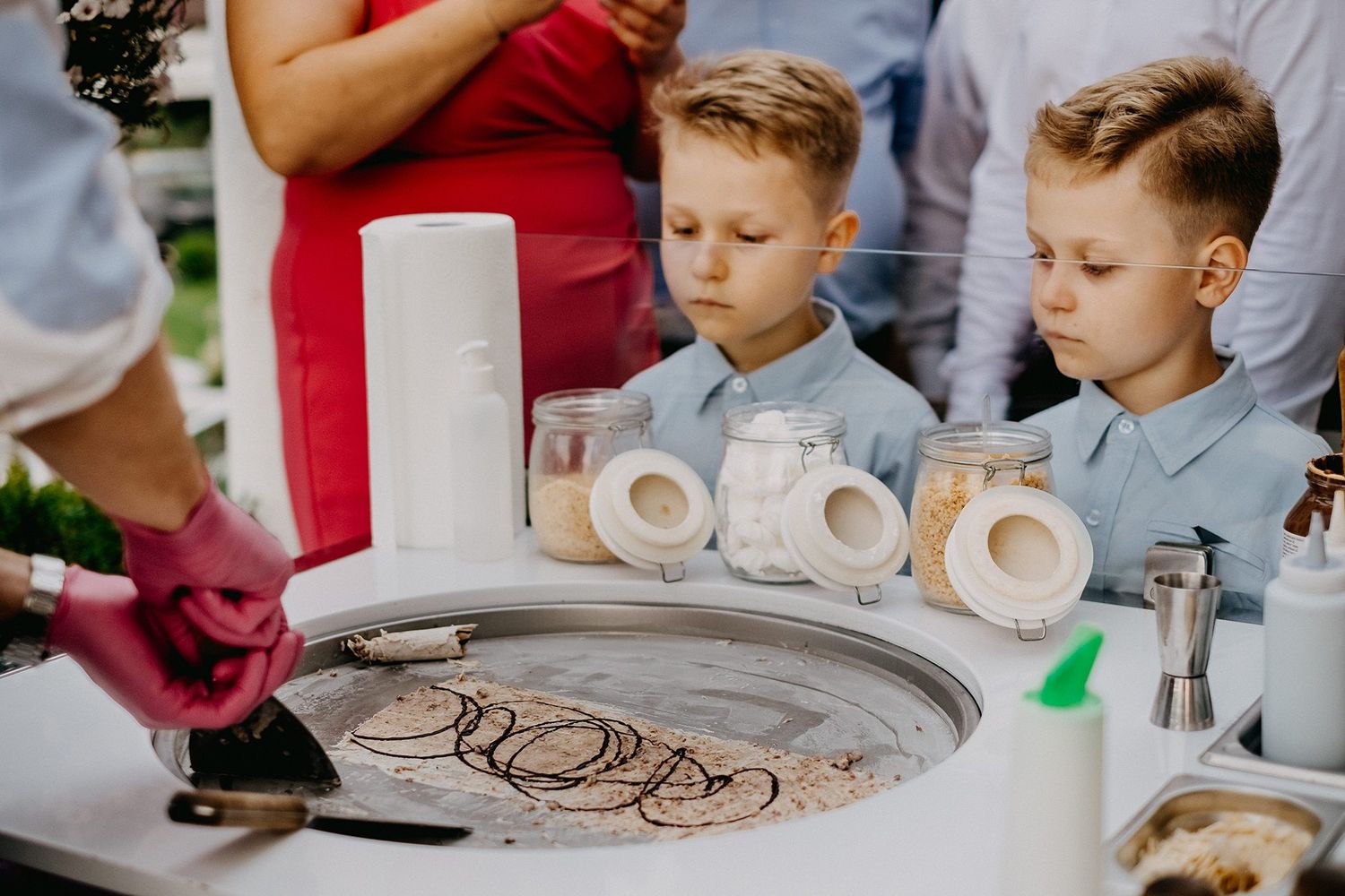 Two young boys in blue shirts watching rolled ice cream being made at an outdoor wedding reception.
