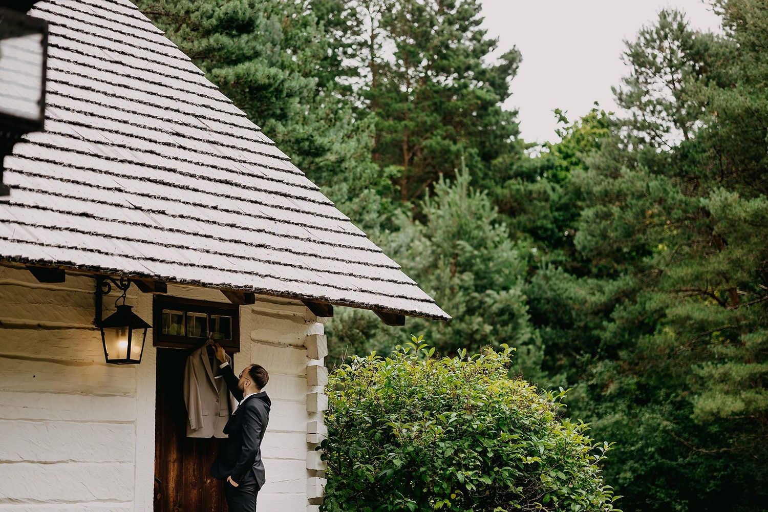 Groom in navy suit standing at entrance of white wooden chapel surrounded by lush green pine trees.