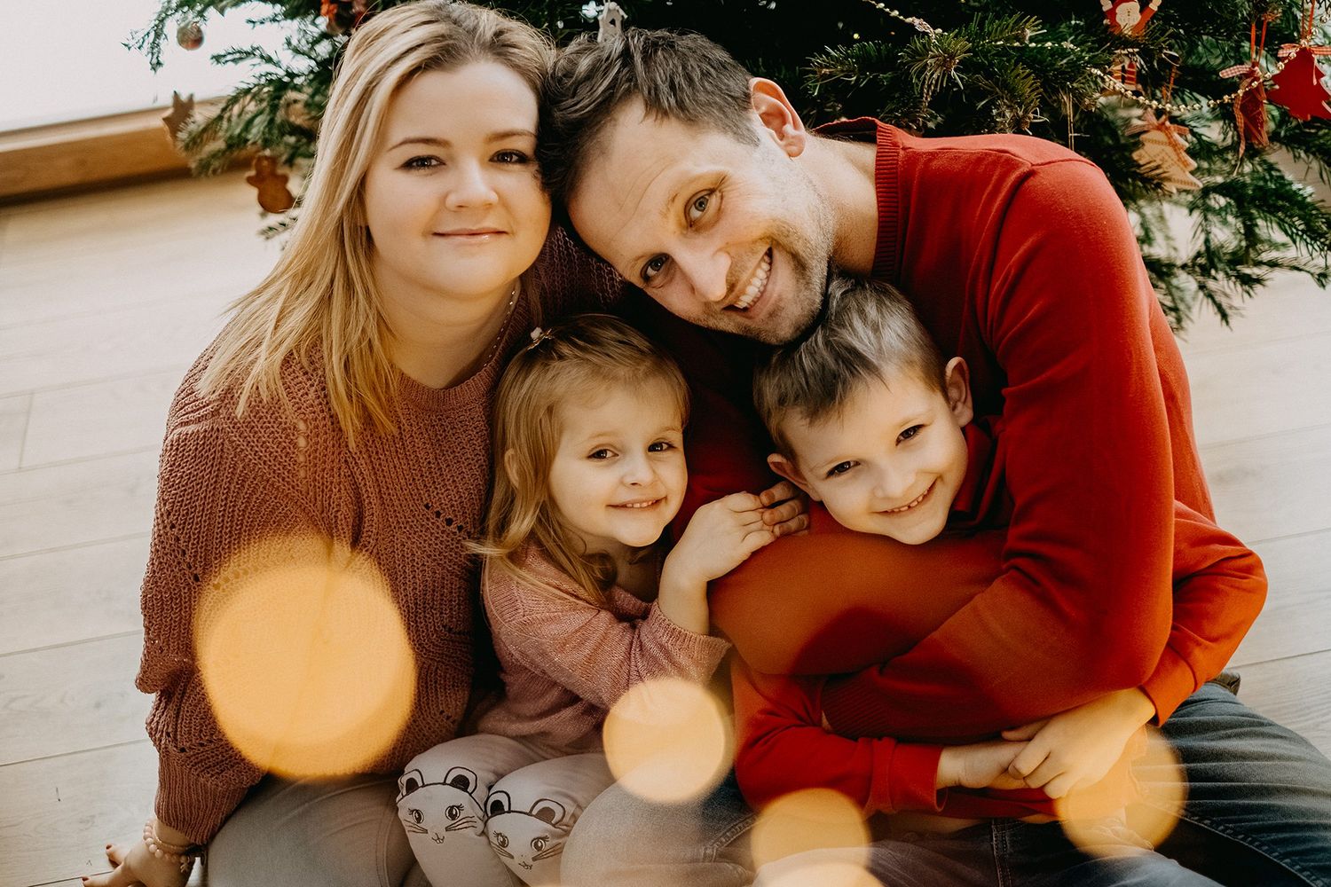 Happy family of four posing together in front of decorated Christmas tree with bokeh lights.