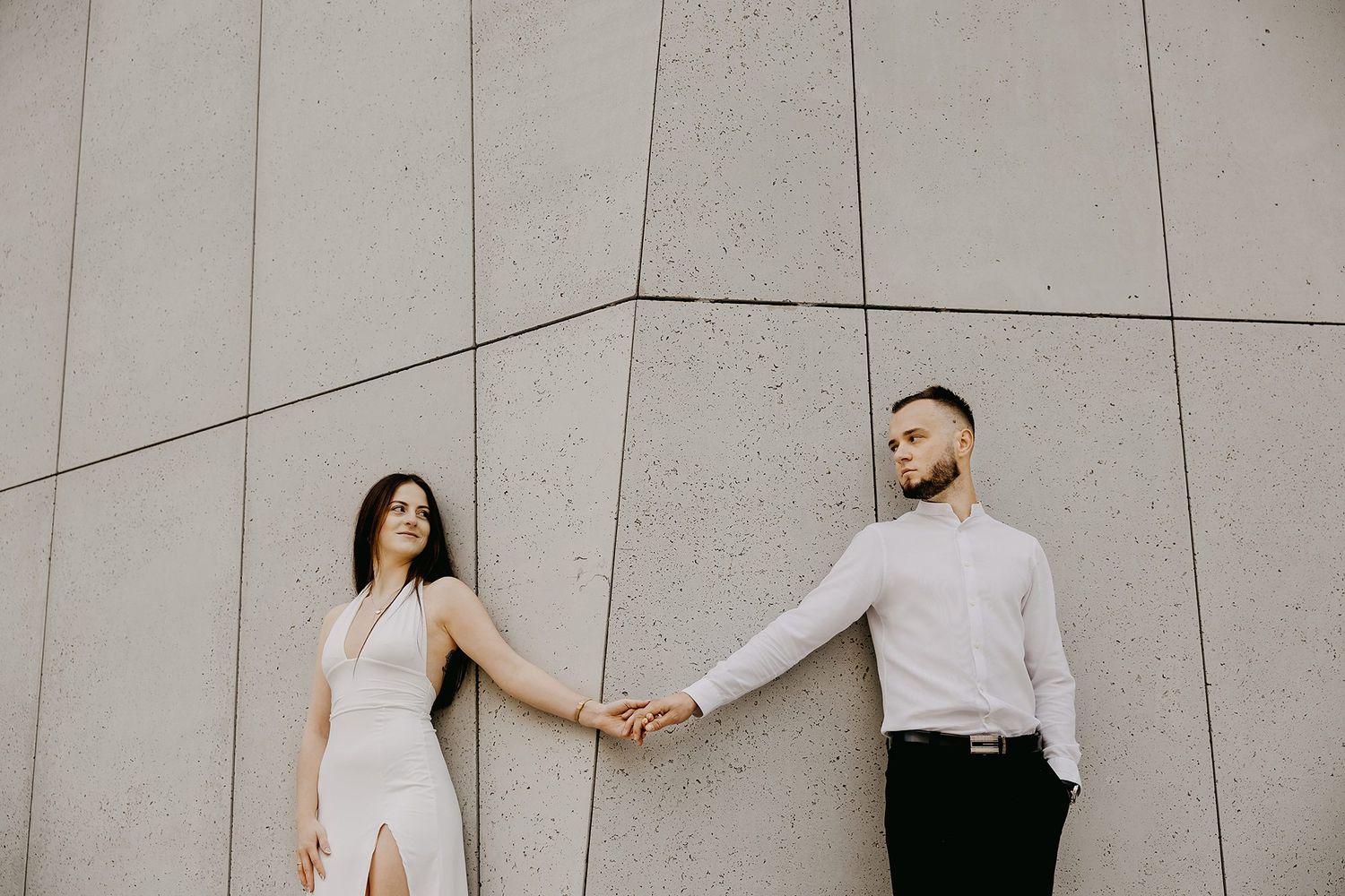 Couple in white outfits holding hands against a modern concrete wall during an elegant engagement photo session.
