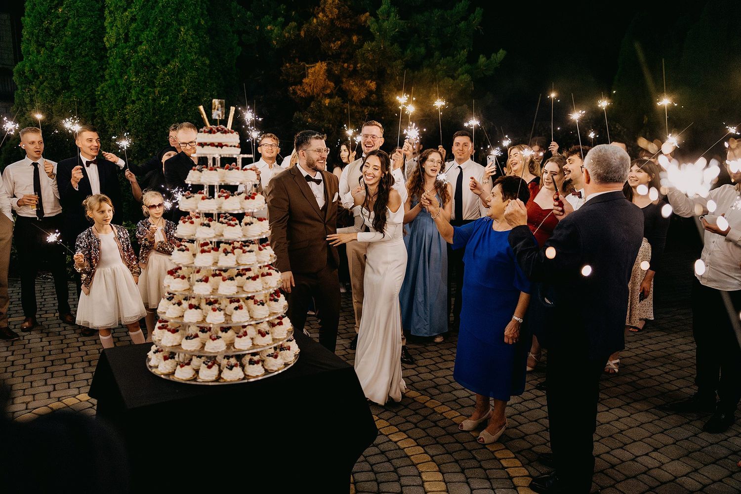 Bride and groom cutting tiered cupcake wedding cake surrounded by guests holding sparklers at night.