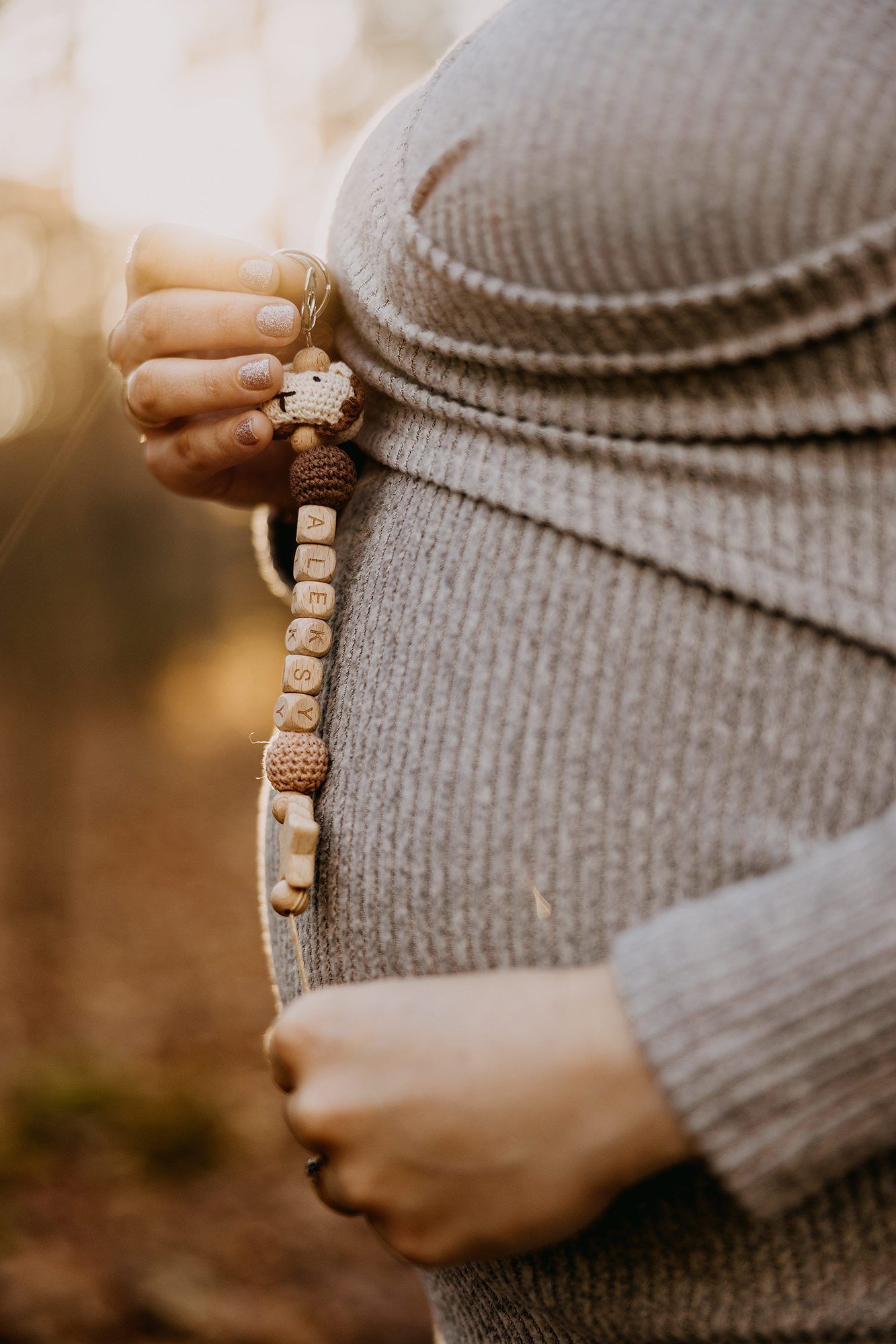 Close-up of pregnant belly holding a personalized wooden bead pacifier clip outdoors.