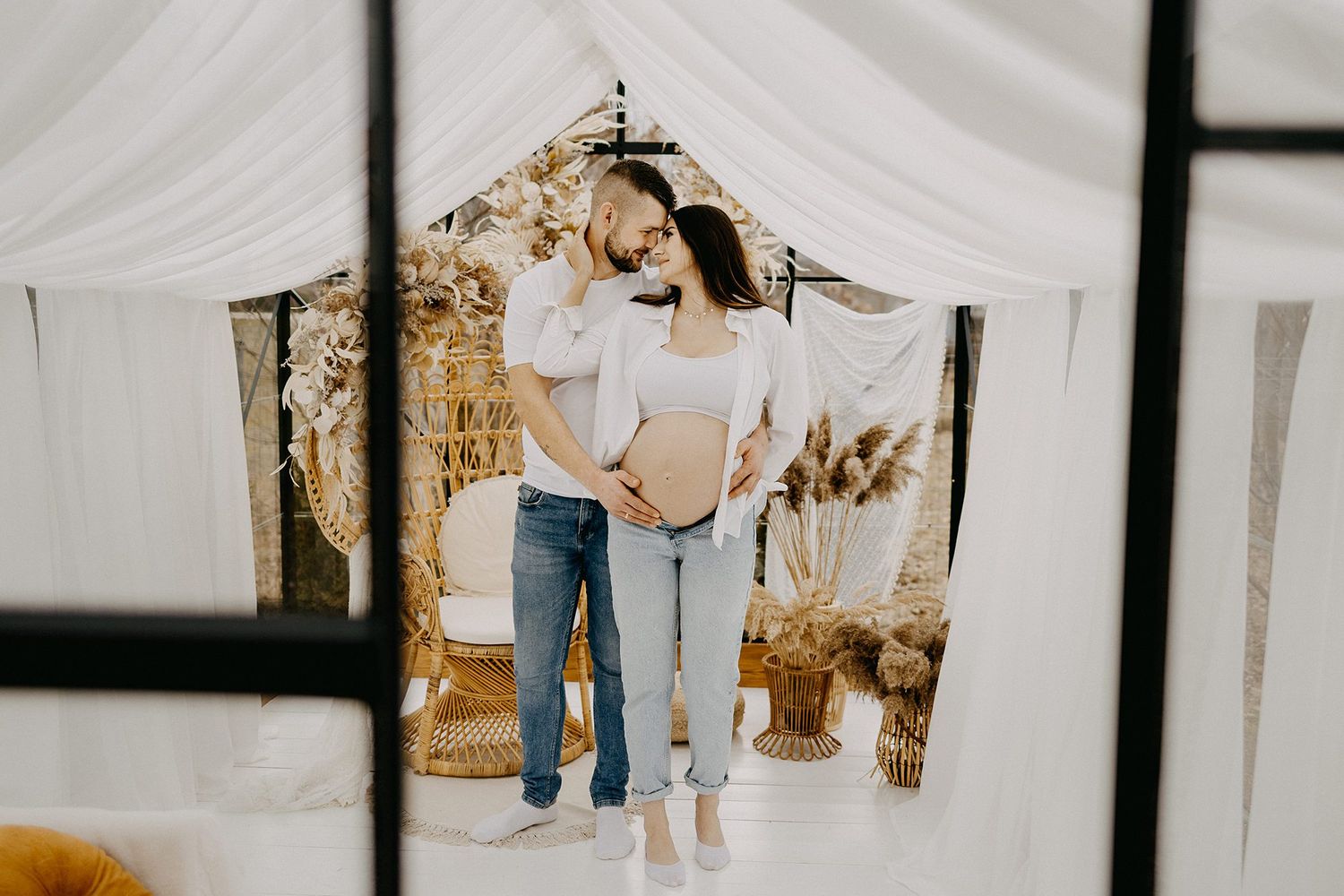 Couple embracing in boho-styled studio with white drapes and dried pampas grass, showcasing pregnancy bump.