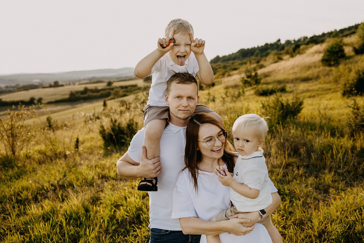 Happy family with two young boys posing in a sunny green field, father holding toddler on shoulders.