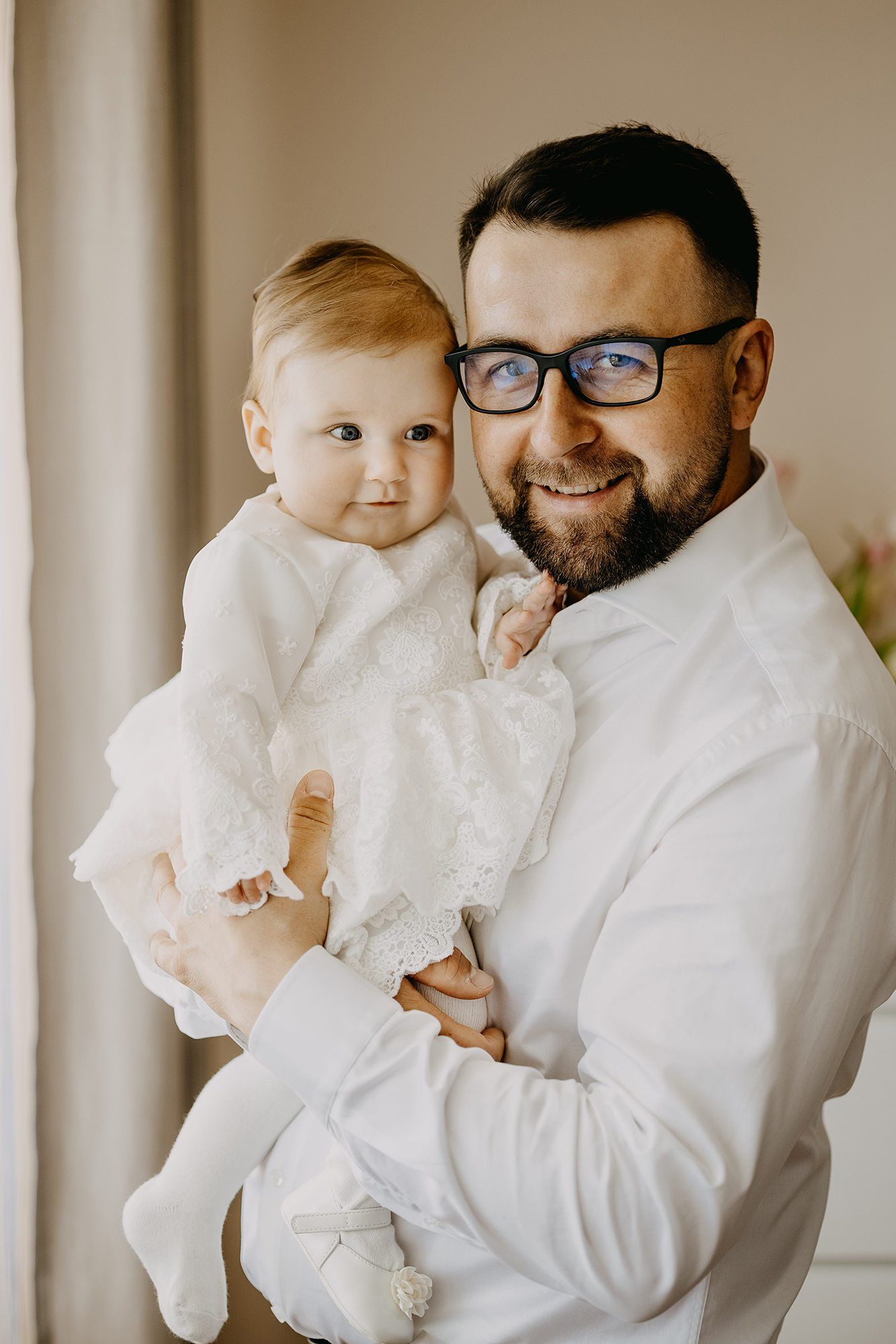 Smiling bearded father wearing glasses holds baby in white lace baptism dress at home.