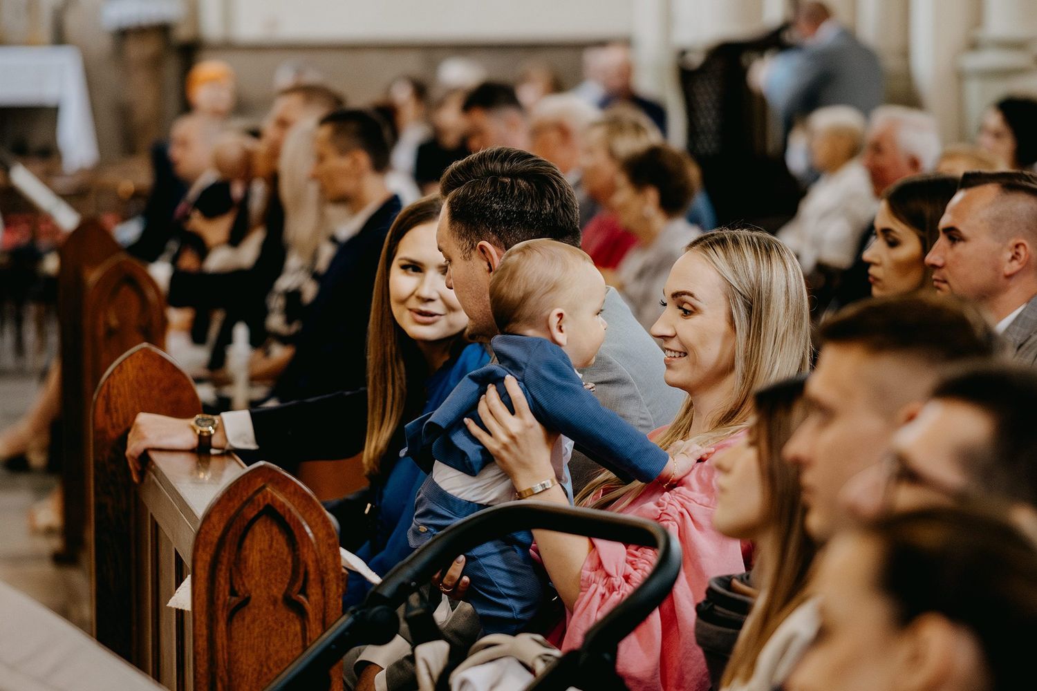 Family with baby sitting in church pew during baptism ceremony surrounded by guests.