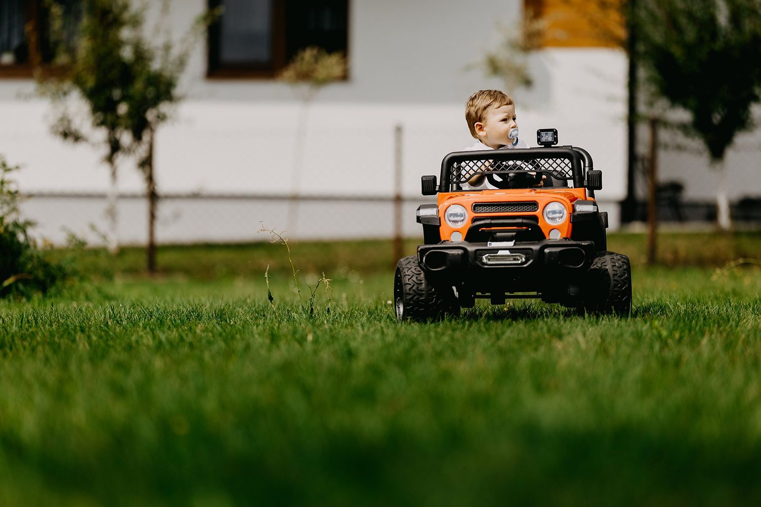 Young toddler boy riding orange electric toy SUV car on green grass in backyard.