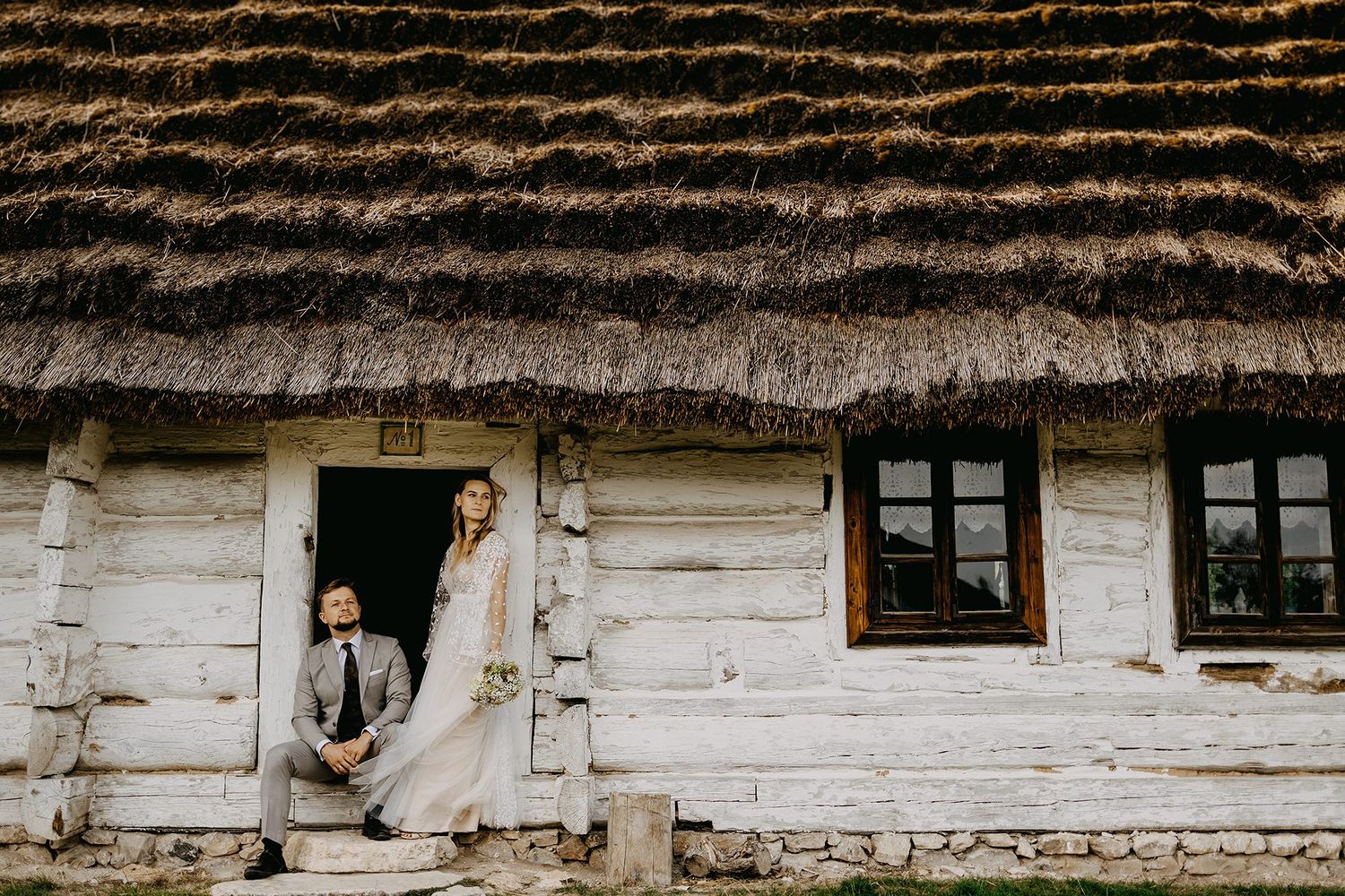 Wedding couple posing at doorway of rustic thatched-roof log cabin in charming countryside setting.