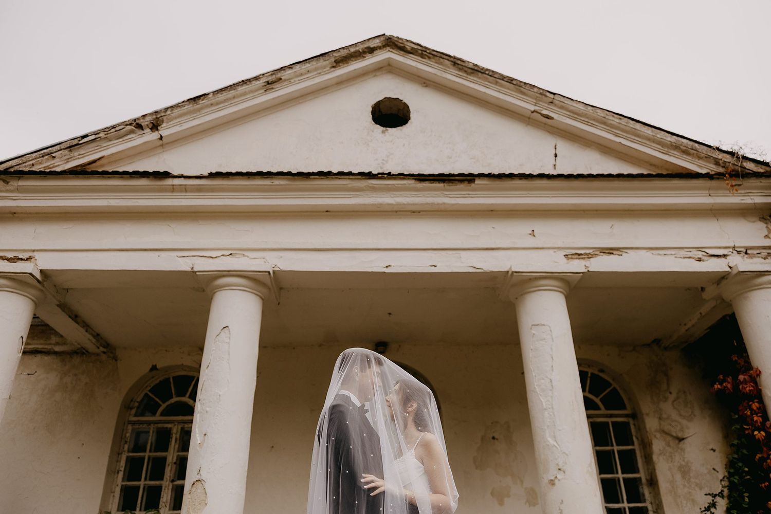 Bride in flowing veil standing before white classical columned building facade.