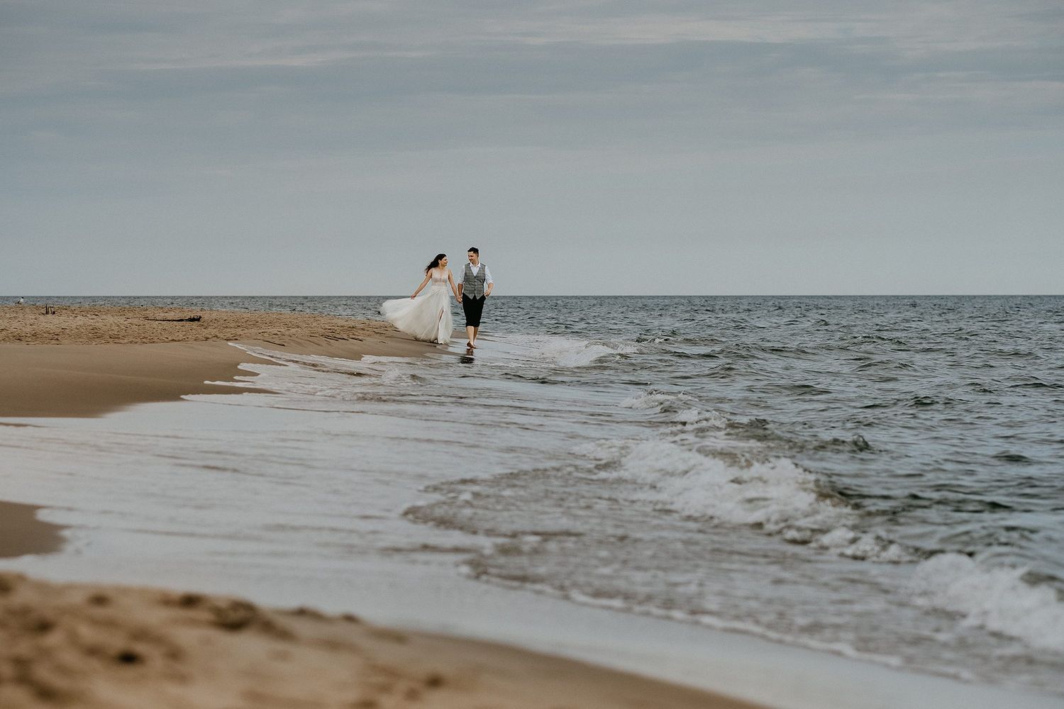 Bride and groom walk along a sandy beach shoreline with gentle ocean waves nearby.