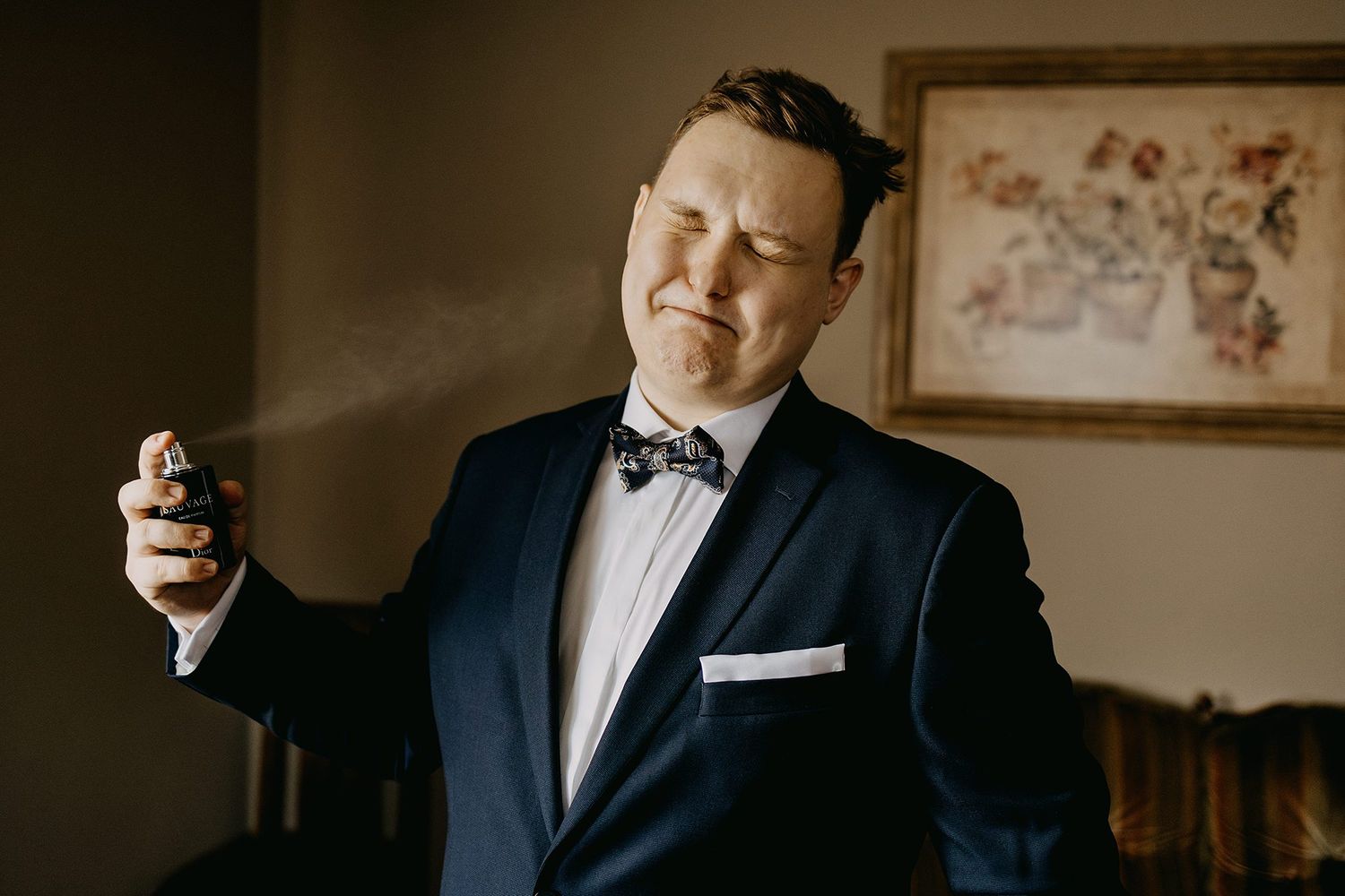 Groom in navy tuxedo spraying cologne with eyes closed before his wedding ceremony.