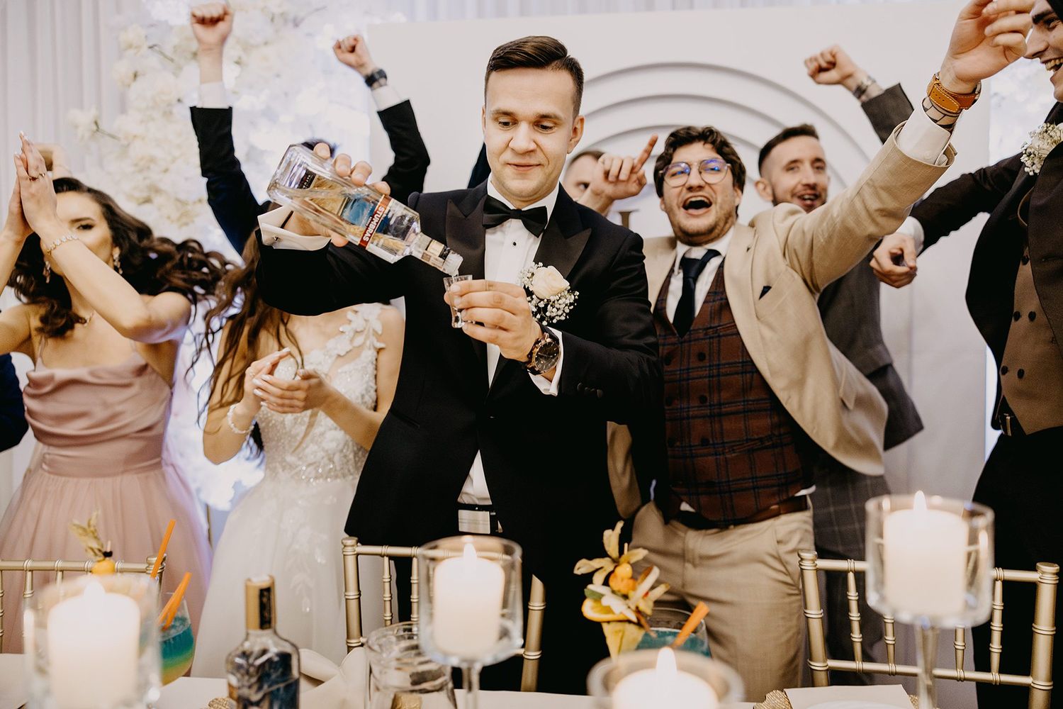 Joyful wedding reception celebration with guests cheering and raising hands around smiling groom in black tuxedo.