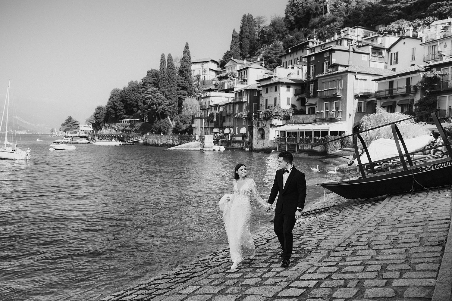 Black and white photo of bride and groom walking hand in hand along Italian lakeside promenade.