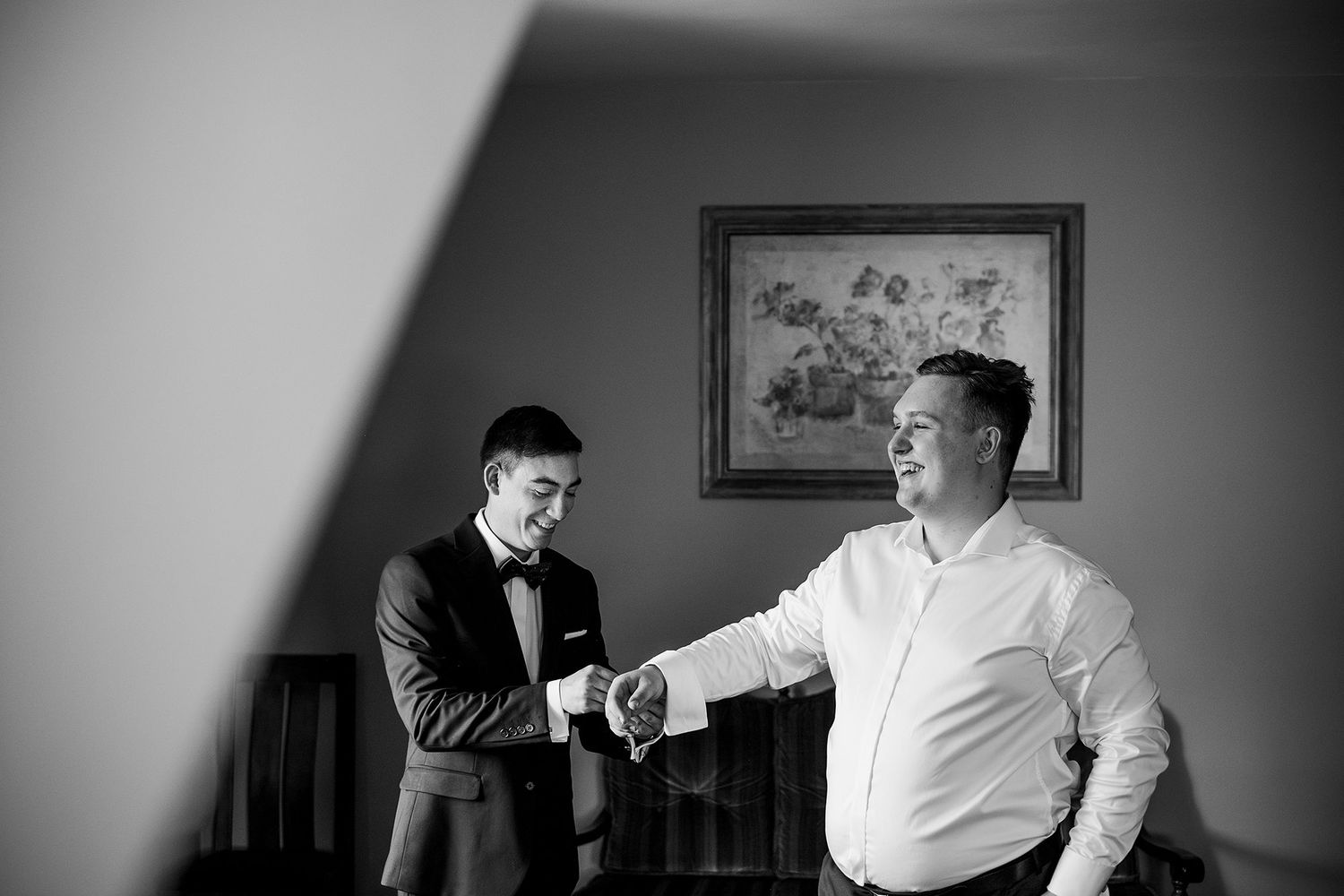 Black and white photo of groom in tuxedo receiving congratulations with a handshake from older man indoors.