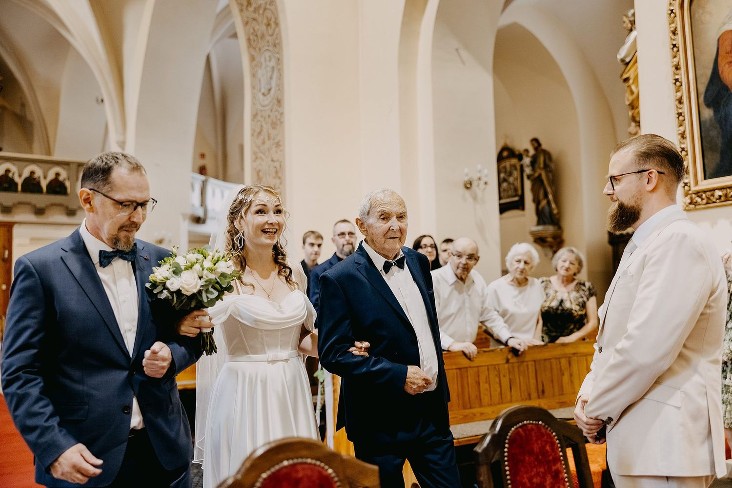 Bride walking down church aisle escorted by two men holding white bouquet.