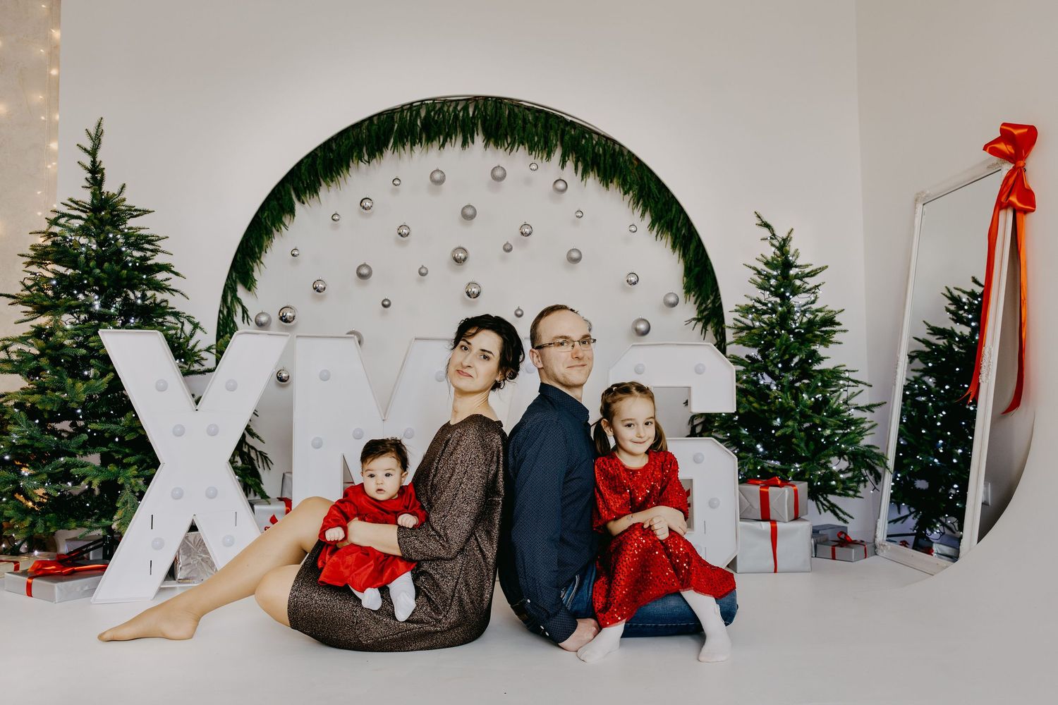 Family of four in red and neutral outfits posing in a bright studio with Christmas trees, gift boxes, and large XO letters.